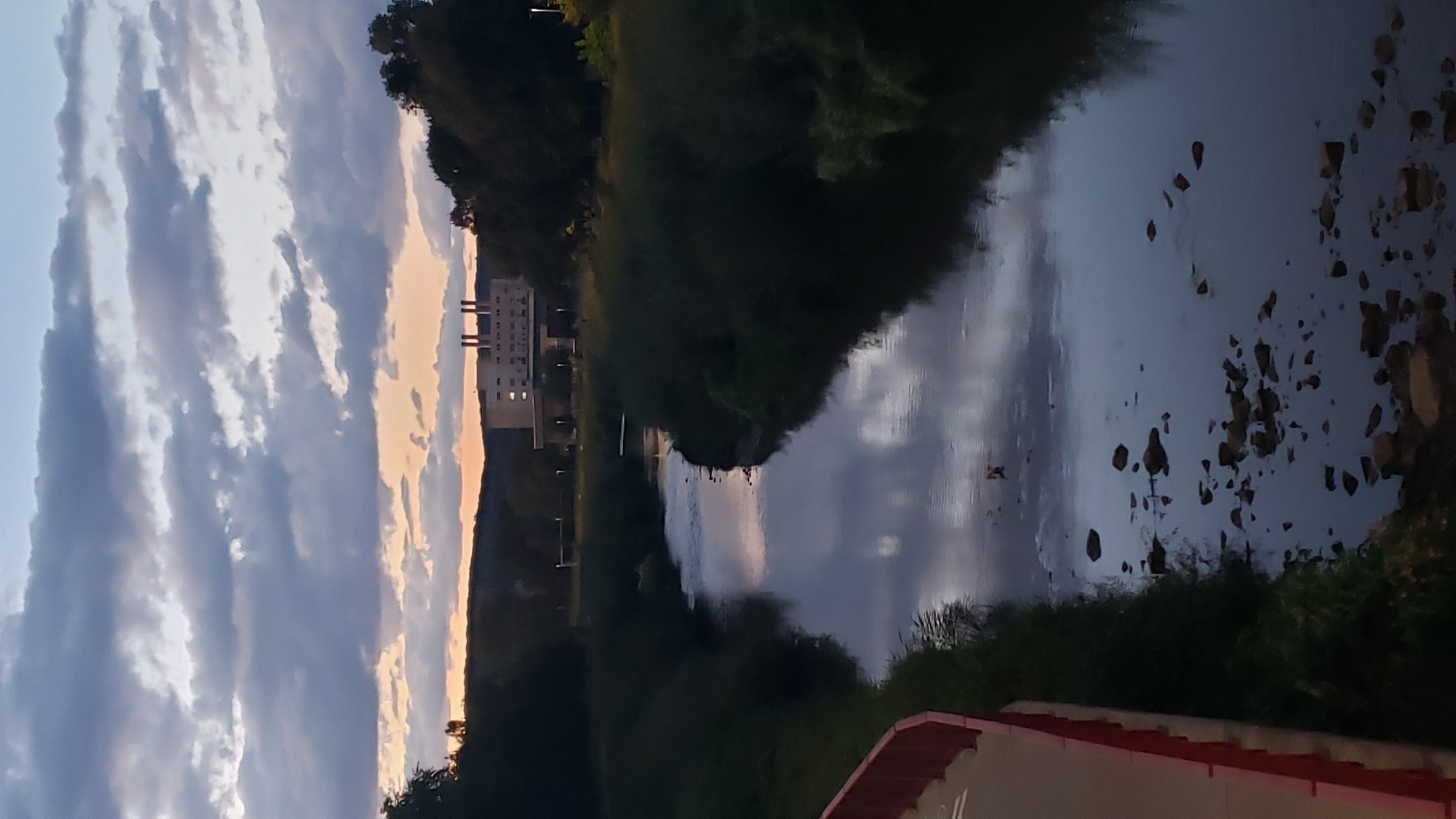 The image depicts a tranquil natural scene at dusk, featuring a winding river reflecting the cloudy sky. The riverbanks are lined with dense, dark greenery, creating a sense of enclosure. In the distance, a multi-story building with several chimneys stands against the backdrop of a hazy, orange-tinged sunset. The foreground shows a red railing, suggesting the viewpoint is from a bridge or elevated walkway. The scene captures a serene moment in what appears to be a semi-urban or suburban environment. The specific location in Prešov, Slovakia, is not identifiable from the visual information provided.