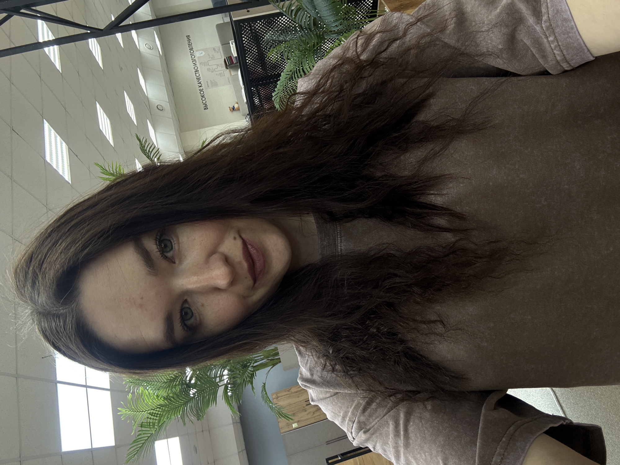 A brunette woman is seated indoors. The photo appears to be taken in a public space with office-like features. She wears a brown, textured shirt. The background includes artificial plants, ceiling lights, and a sign in Russian, which references quality manufacturing. This potentially places the location in an industrial or commercial area of Chaikovsky, Russia.
