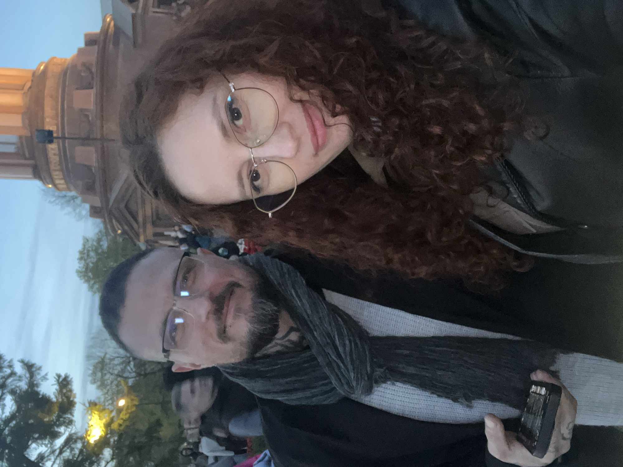 Two people pose for a selfie. A woman with long curly brown hair and round glasses is in the upper part of the frame. A man with a beard, glasses, and a grey scarf is in the lower part of the frame. A building with a dome is visible behind them. The photo appears to be taken in a public area, possibly a square or park, during the evening or dusk. There are trees and other people in the background. The man is holding a small black rectangular object, possibly a phone or a remote. Based on the architecture of the building in the background, the location is likely the Palace Square in Saint Petersburg, Russia, with the General Staff Building visible.