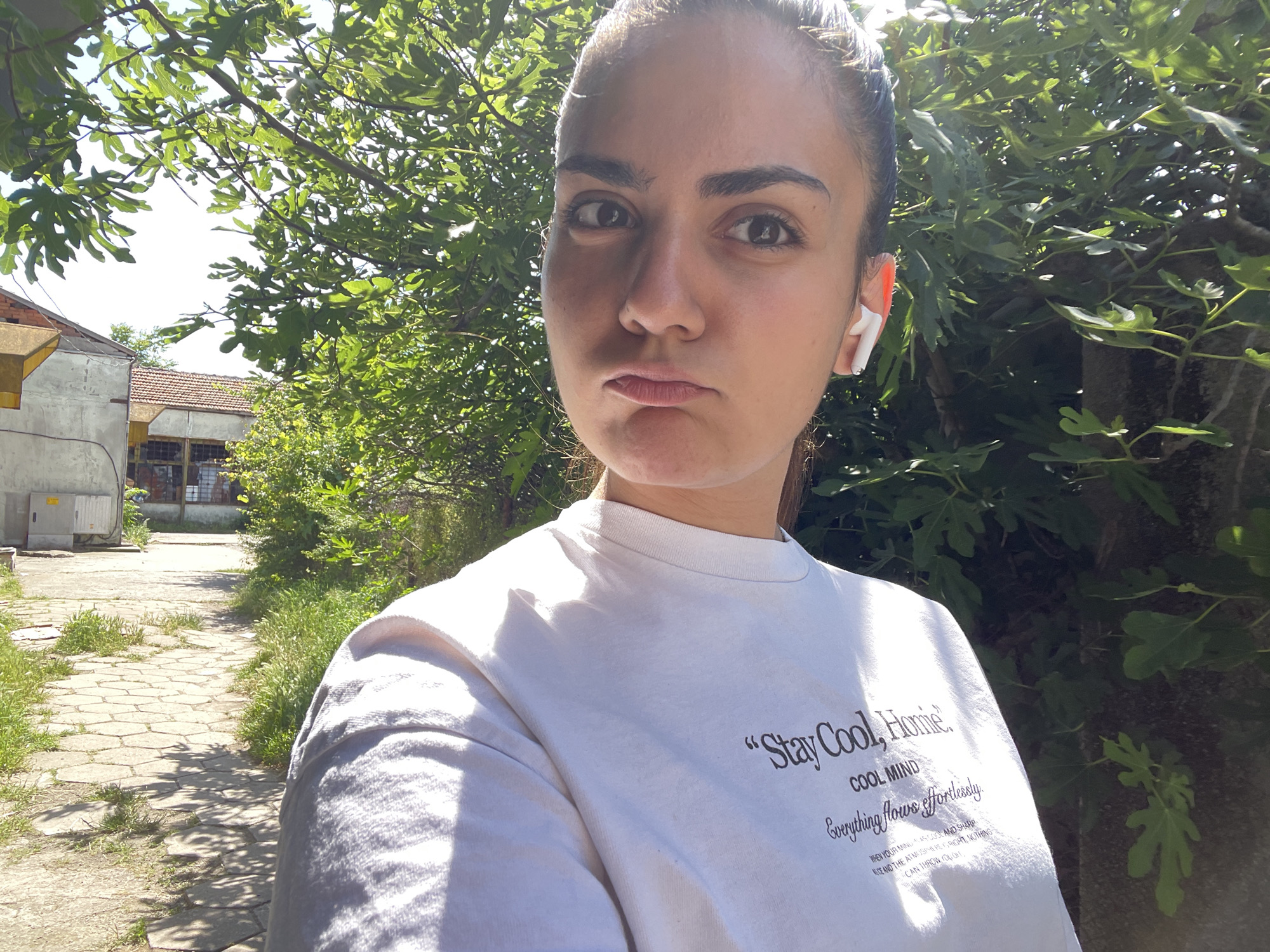 A young woman with dark hair pulled back, wearing a white t-shirt with text, is taking a selfie outdoors. She has on white wireless earbuds and is looking at the camera with a neutral expression. The background shows lush green foliage, a paved walkway, and some buildings. The scene is illuminated by bright sunlight, casting shadows. The location is identified as Debelt, Bulgaria.