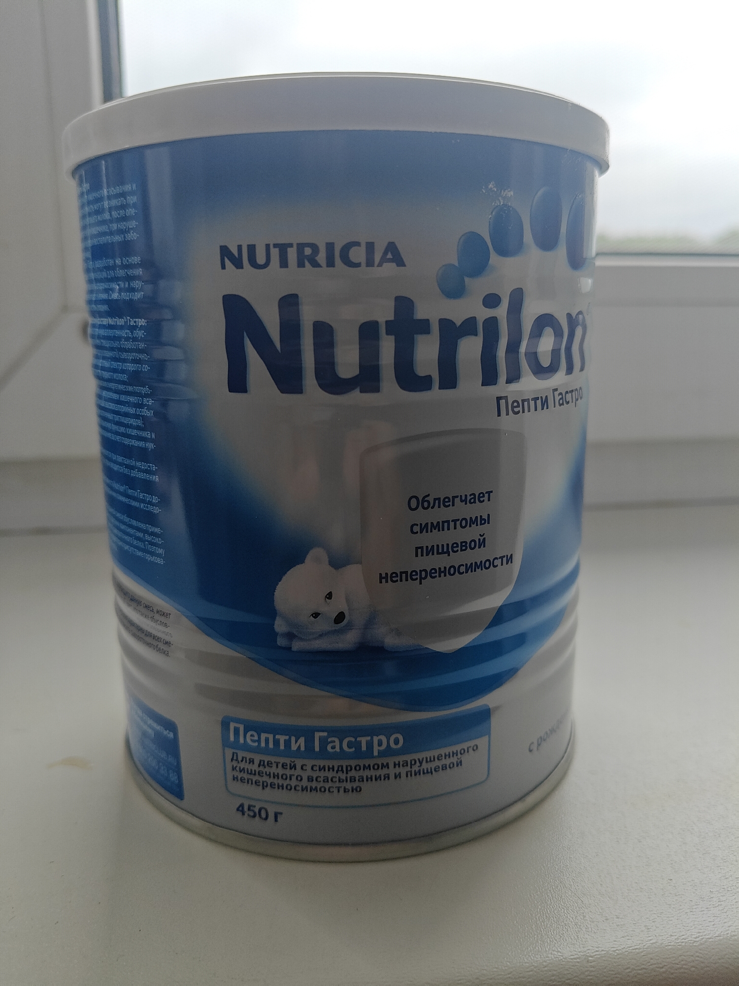 The image shows a white and blue can of Nutricia Nutrilon Pepti Gastro formula, with a label that includes text in Russian. The can is positioned on a white surface, likely a windowsill, with a blurred background that suggests an outdoor environment with overcast skies. The can is the primary subject of the image. There are no people or animals visible in the image. The lighting is natural and diffused. The image appears to have been taken in an indoor setting that overlooks an outdoor area. The presence of the Nutricia brand suggests a commercial product.