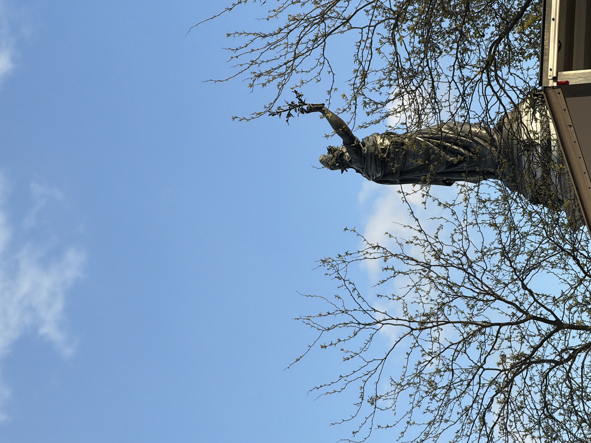 A low-angle shot of the statue of “La Gloire de l’Armée Française” (Glory of the French Army) at the Pont d'Iéna in Paris, France, taken during the day. The copper statue is positioned against a blue sky with scattered clouds. A tree with bare branches is partially obscuring the statue and the sky. The statue is mounted atop a stone pedestal, which is partially hidden in the frame. The statue is draped in robes, and its right arm is raised, holding what appears to be a laurel branch. Part of the statue is next to the side of what appears to be a white truck. The photograph is rotated counterclockwise.