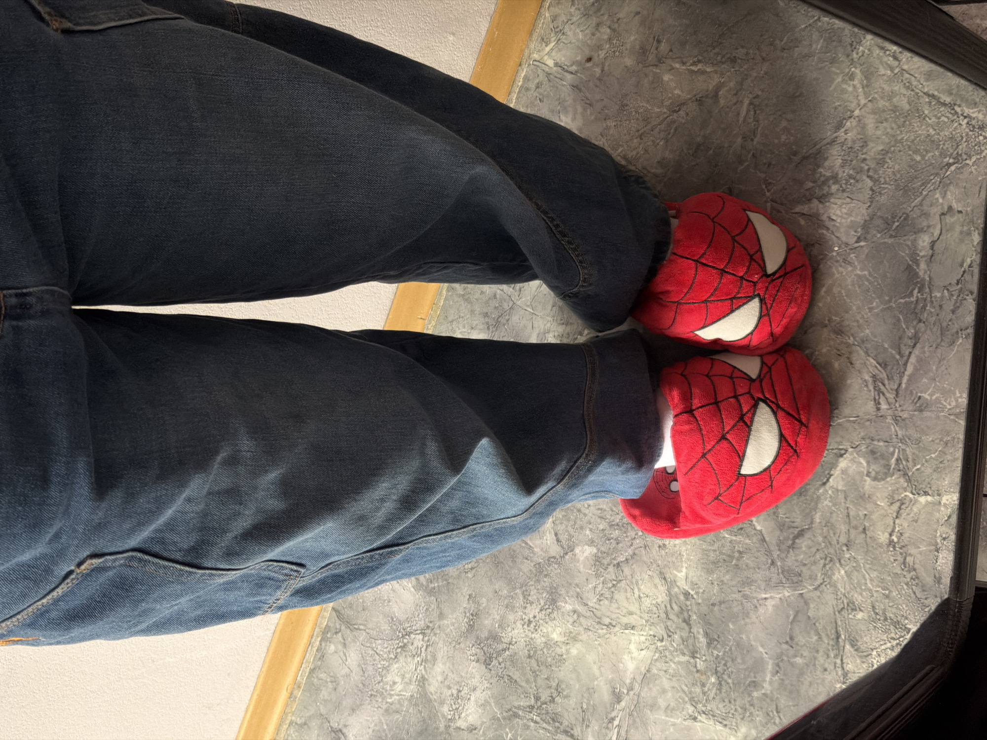 A top-down view shows a person wearing blue jeans and red Spider-Man themed slippers. The person is indoors, standing on a gray and white marbled tile floor. A wooden trim is visible along the edge of the floor. A corner of a black object is in the upper right corner. The image does not include information about Perm, Russia.