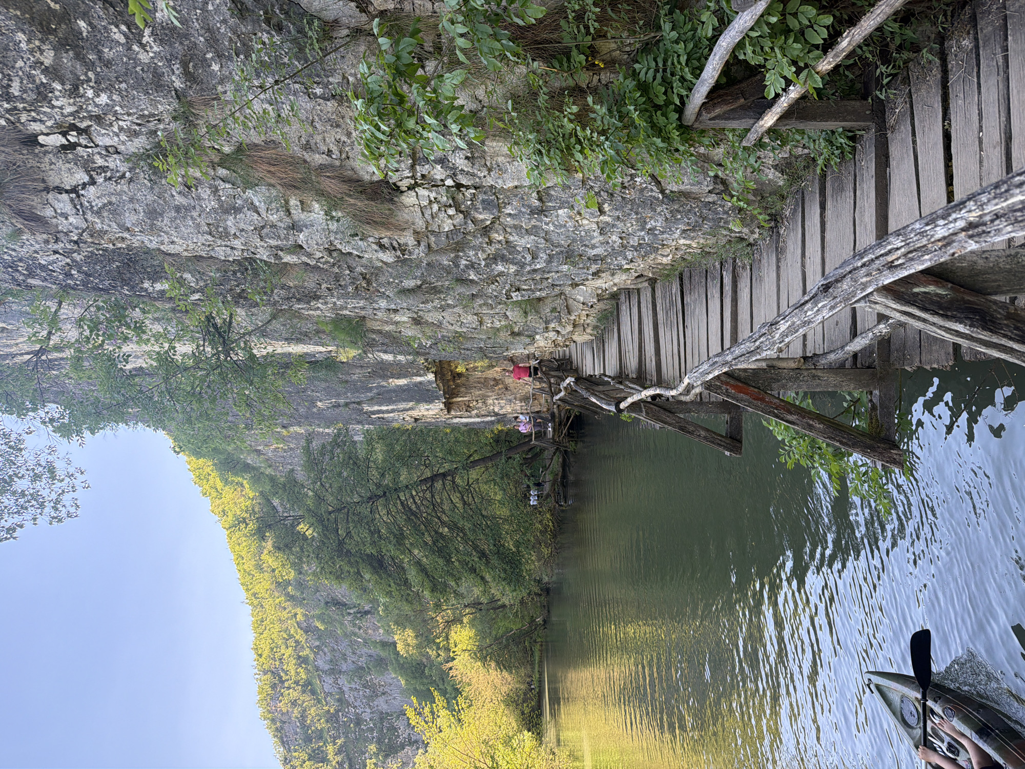 The image shows a view from the Iskar Panega Geopark near Petrevene, Bulgaria. A wooden walkway clings to a rocky cliff face. The walkway extends over a body of water, its surface reflecting the cliff and surrounding vegetation. People are seen on the walkway in the distance. In the foreground, a kayak with a paddle is partially visible. The sky is blue. The scene is dominated by the natural elements: rock, water, and vegetation.