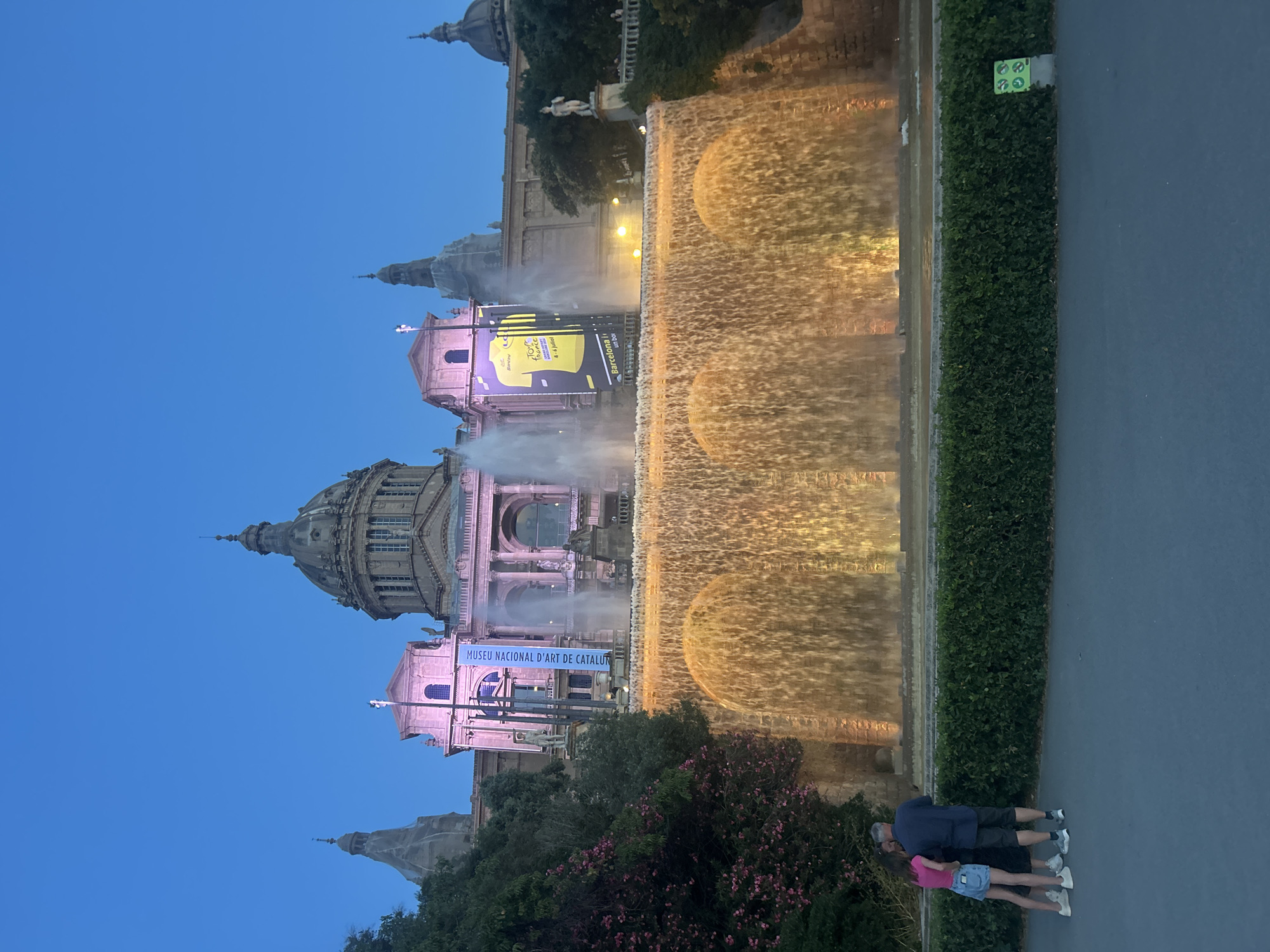 The image shows the Magic Fountain of Montjuïc in Barcelona, Spain, at dusk. A large, ornate building, which is the National Museum of Art of Catalonia, stands prominently in the background. The fountain itself is the main subject, with water cascading down multiple tiers, illuminated by warm, golden lights. Large jets of water are also spraying upwards in front of the building.
In the foreground, three people are standing near a low, manicured hedge. Two women are on the left, one in a pink top and denim skirt, the other in a dark dress. A man in a dark blue shirt and shorts stands between them, with his arm around one of the women. They appear to be looking at the fountain.
A large banner hangs from the building, displaying a yellow and black image of a cycling jersey with the text "Le Tour de France" and "Barcelona un pel". The sky is a deep twilight blue, indicating the transition from day to night. The overall atmosphere is one of a public gathering place, showcasing a significant architectural landmark and its associated light and water show.