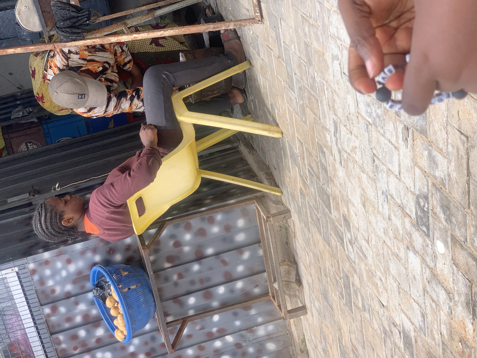 In Dadi Riba, Nigeria, a young woman with braided hair sits on a yellow plastic chair, looking to her right. She wears a maroon sweater and dark pants. In front of her, to her left, a blue basket overflows with potatoes on a metal shelf. Behind her, another person, wearing a cap and a colorful patterned dress, bends over. The background consists of a corrugated metal wall and a paved courtyard. A hand in the foreground is holding a beaded bracelet.