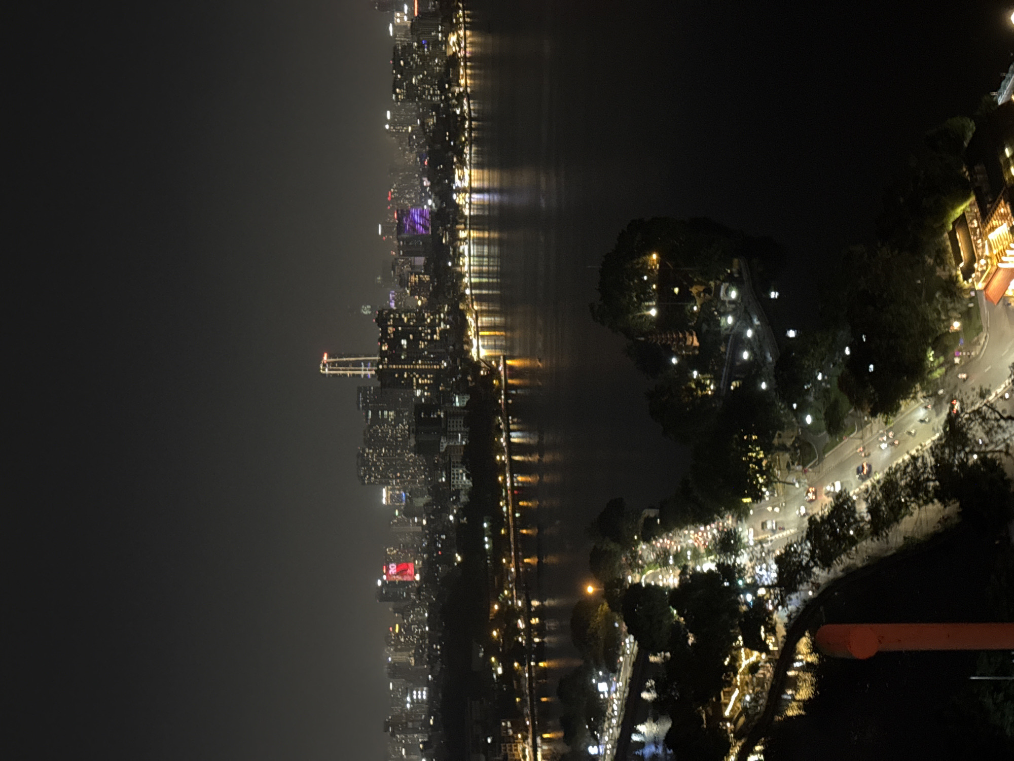 This is a nighttime aerial view of Hoan Kiem Lake in Hanoi, Vietnam. The lake's dark water reflects the lights of the surrounding cityscape, which includes numerous illuminated buildings and skyscrapers in the distance. A tree-lined street with traffic is visible along the edge of the lake in the foreground, with lights from street lamps, businesses, and passing vehicles creating a vibrant glow. On a small island in the lake, a pagoda is visible, illuminated by lights. The overall impression is of a bustling urban environment at night, with the lake serving as a central point of reflection and tranquility within the city.