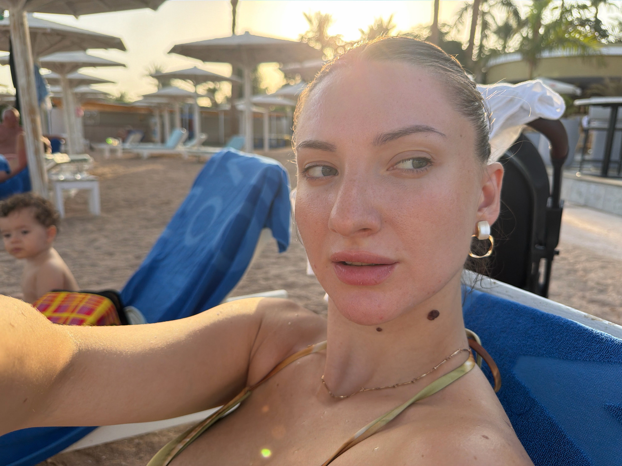 A woman, partially visible, is seen in a selfie at a beach resort in Sharm El Sheikh, Egypt. She is wearing a bikini top and a necklace. Her face is turned to the side, and she is looking away from the camera. In the background, beach chairs and umbrellas are visible, along with palm trees and the sea. A child can be seen in the background, to the left of the woman. The lighting suggests it is late afternoon or early evening, with the sun casting a warm glow over the scene. The woman has several moles on her face and neck. She is sitting on a blue beach chair with a towel draped over it.