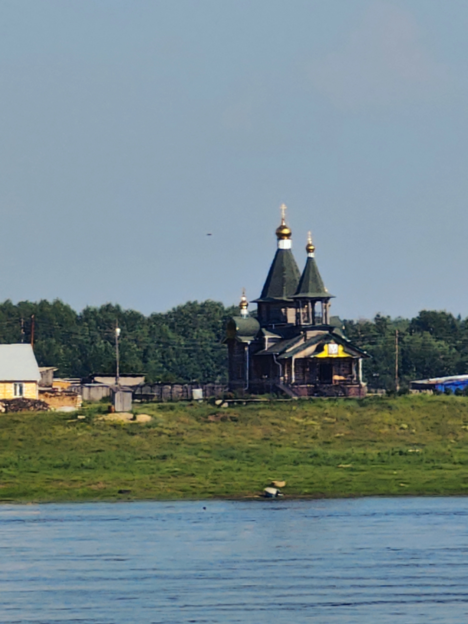 The image depicts a serene landscape with a prominent wooden church featuring green roofs and golden domes situated on a grassy hill. The church is surrounded by a few other buildings, including a yellow house with a white roof and what appears to be a small shed. A body of water, likely a river, occupies the foreground, reflecting the blue sky. In the background, a dense forest line stretches across the horizon under a clear, pale blue sky. A small dark object, possibly a bird or a drone, is visible in the sky. The overall scene suggests a peaceful rural setting in Потапово, Russia.