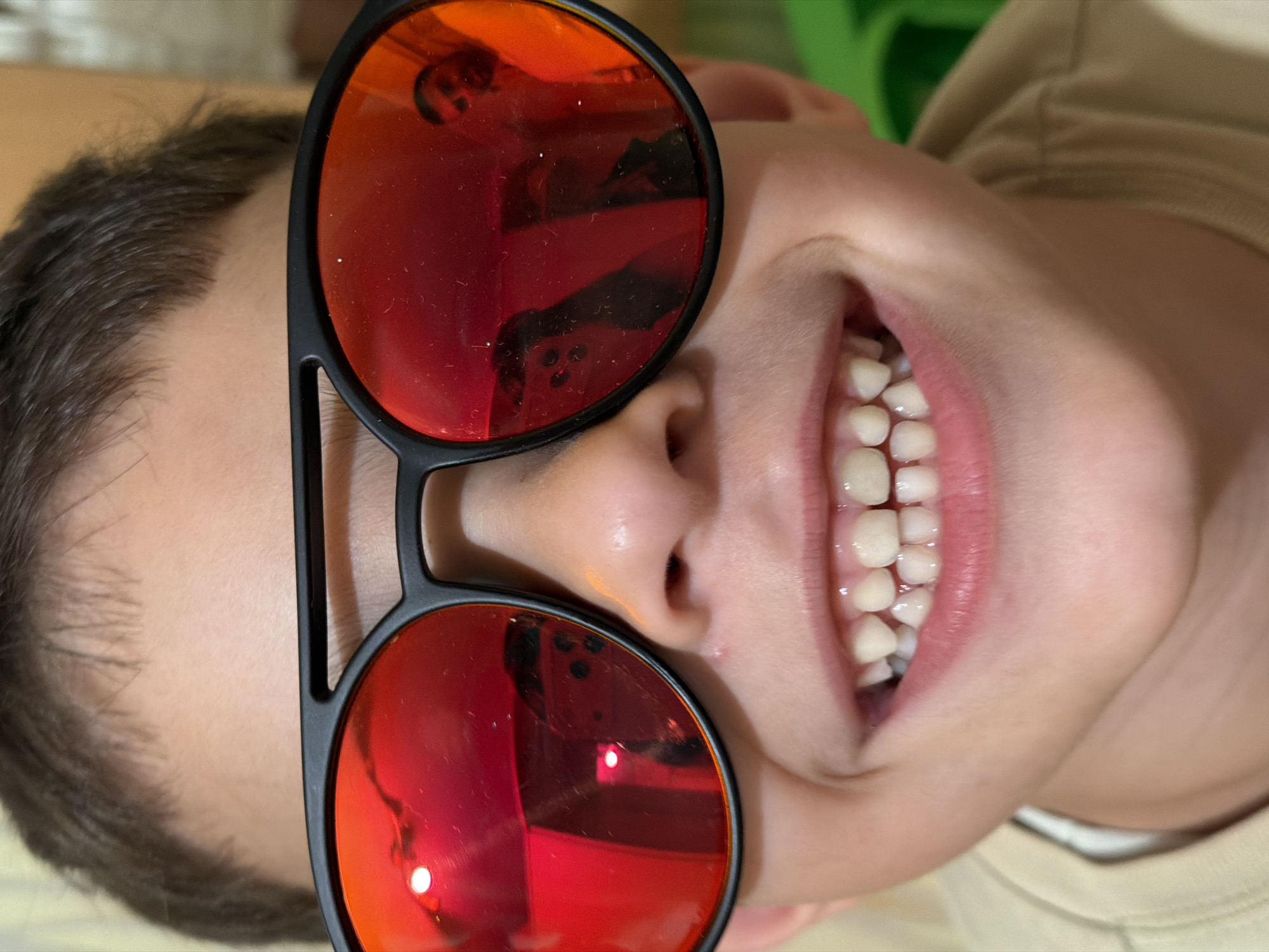 A close-up, head-on shot captures a young child wearing oversized red-tinted sunglasses. The child's mouth is open in a wide smile, revealing a set of healthy, white teeth. The sunglasses have thick black frames with a double bridge. Reflections in the lenses show a person with a beard and dark hair. The background is softly blurred, with hints of green on the right side and a beige wall or fabric on the left. The child's short brown hair is visible above the sunglasses. The lighting is bright and appears to be coming from the front, creating reflections and highlights on the sunglasses and skin.