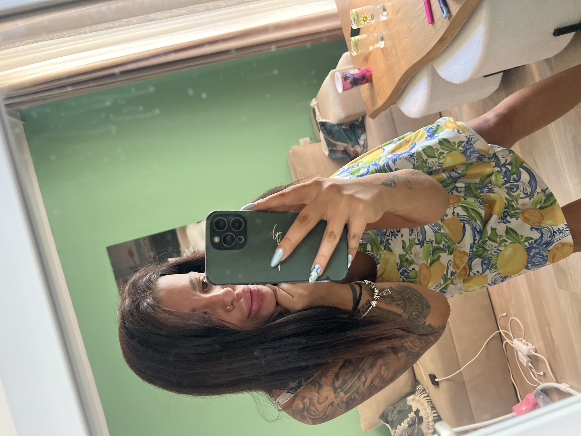 A young woman with dark hair and tattoos on her left arm takes a selfie in a mirror. She is wearing a yellow and blue patterned dress. She is leaning on her hand and looking at the camera. In the background, there is a couch, a table with drinks, and a green wall. The scene appears to be indoors, possibly in a home. There is no identifiable information that would allow for the determination of a specific location within the city of Plovdiv, Bulgaria.