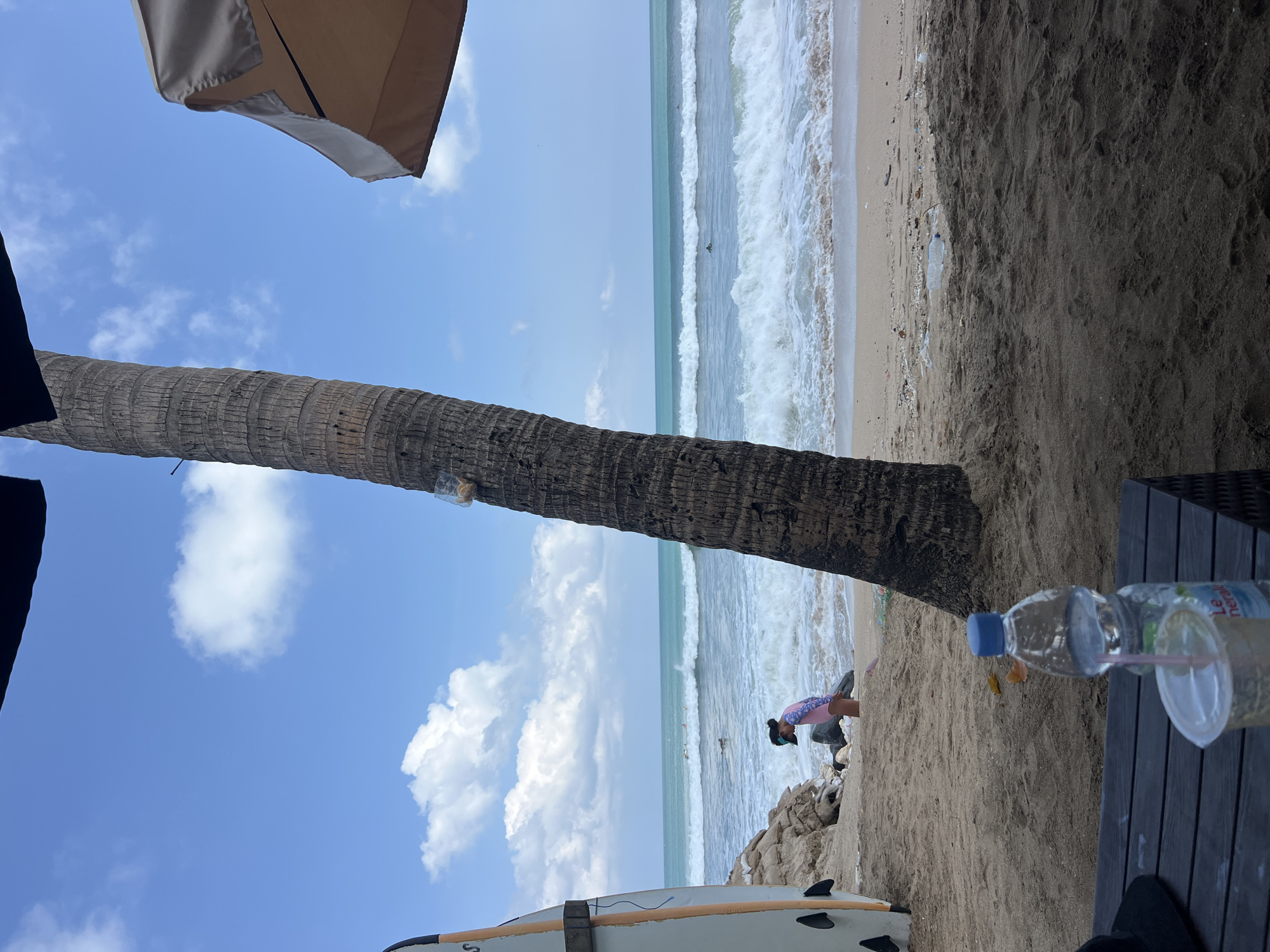 The image depicts a beach scene in Kuta, Indonesia. In the foreground, a dark wooden table is visible, holding a partially consumed plastic cup with a straw and a bottle of water. To the left of the table, a black hat rests. Behind the table, a person is partially visible, with their head and shoulders showing.
Further back, a large palm tree trunk dominates the mid-ground, with a plastic bag tied to it near the top. To the left of the palm tree, a white surfboard stands upright, leaning against some rocks.
In the background, a person wearing a colorful swimsuit and a dark visor is standing near the shore, facing the ocean. The ocean itself is active, with moderate waves breaking on the sandy beach. Several surfers are visible in the water further out. The sky is blue with scattered white clouds. The beach shows signs of litter, including plastic bottles and debris. A beach umbrella is partially visible at the top right of the image.
