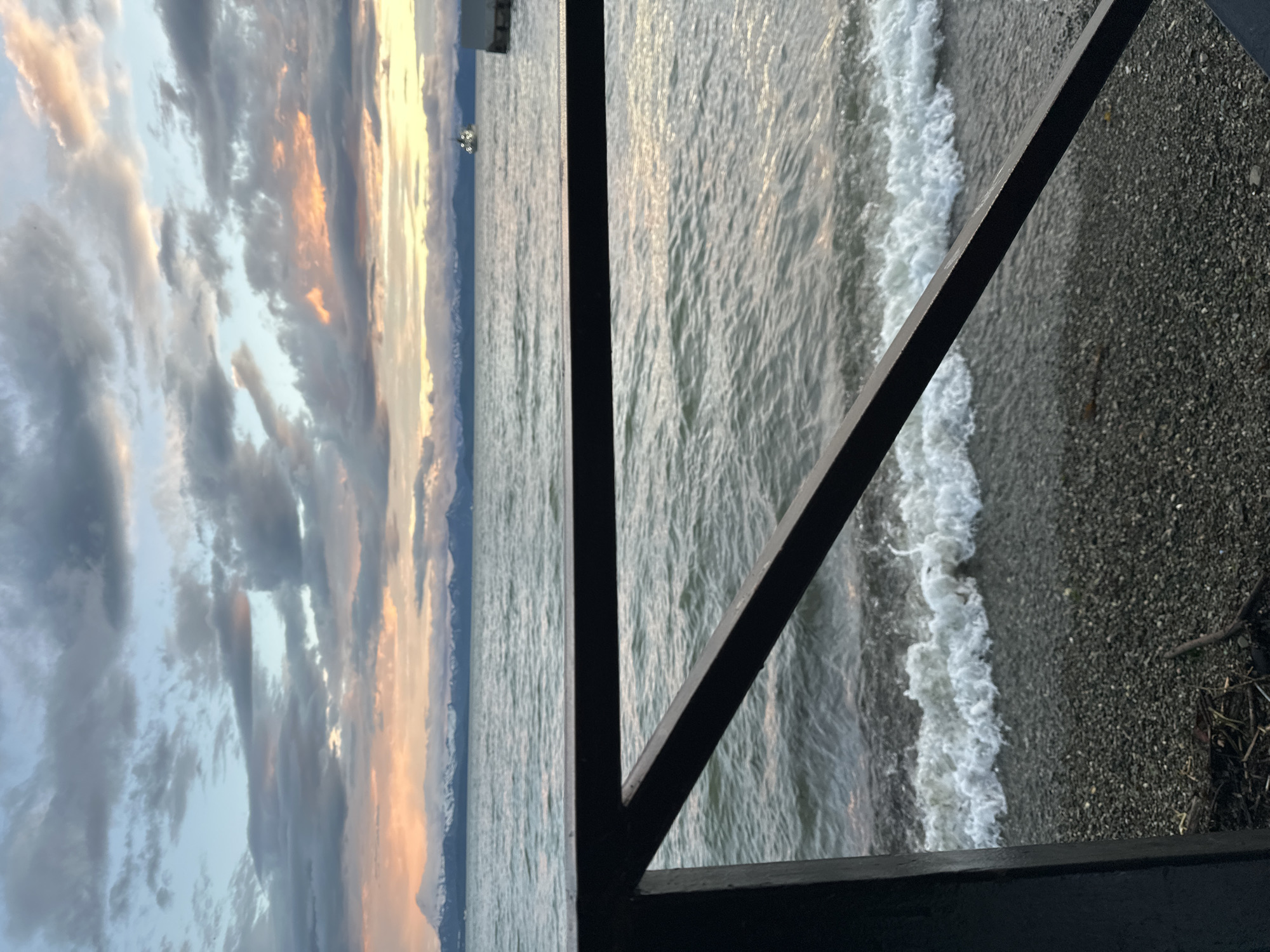 The image depicts a serene waterscape with a dramatic sky at either dawn or dusk. In the foreground, a dark, angular railing partially obscures the view of a pebble beach and the gently lapping waves of a body of water. The water is a deep grey-green, reflecting the subdued colors of the sky. Beyond the water, a distant mountain range with snow-capped peaks forms a majestic backdrop. The sky is a canvas of swirling clouds, with soft hues of pink, orange, and yellow mingling with shades of grey and blue, suggesting the presence of either sunrise or sunset. In the far distance, a small boat with lights on is visible on the water, adding a sense of scale and hinting at human activity. The overall atmosphere is one of tranquility and natural beauty. Based on the mountainous terrain and the presence of a large body of water, this scene is likely located near Petropavlovsk-Kamchatskiy, Russia, possibly overlooking Avacha Bay, with the Vilyuchinsky volcano or other peaks of the Kamchatka peninsula visible in the distance.