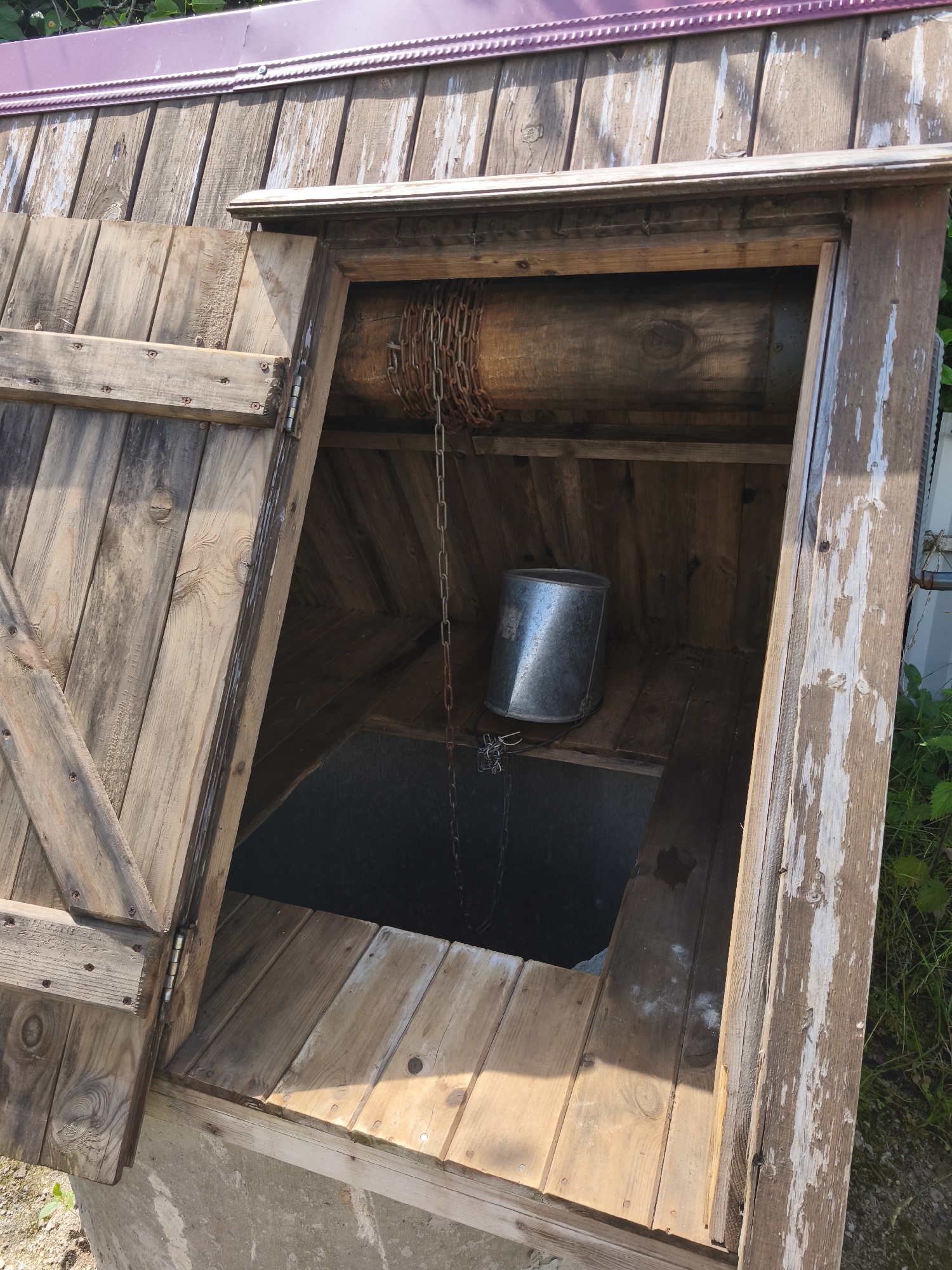 This is a close-up, eye-level, outdoor, medium shot of a rustic wooden well structure. The well has a pitched roof made of purple metal sheeting. A wooden door on the left side is open, revealing the interior. Inside the well, a metal bucket hangs from a chain attached to a wooden roller mechanism. The wooden structure appears weathered and aged, with visible screws and some peeling paint. The well is situated in a natural environment with green foliage visible in the background and to the right. The ground around the base of the well appears to be a mix of dirt and gravel. The image was taken in Dolzhikovo, Russia.