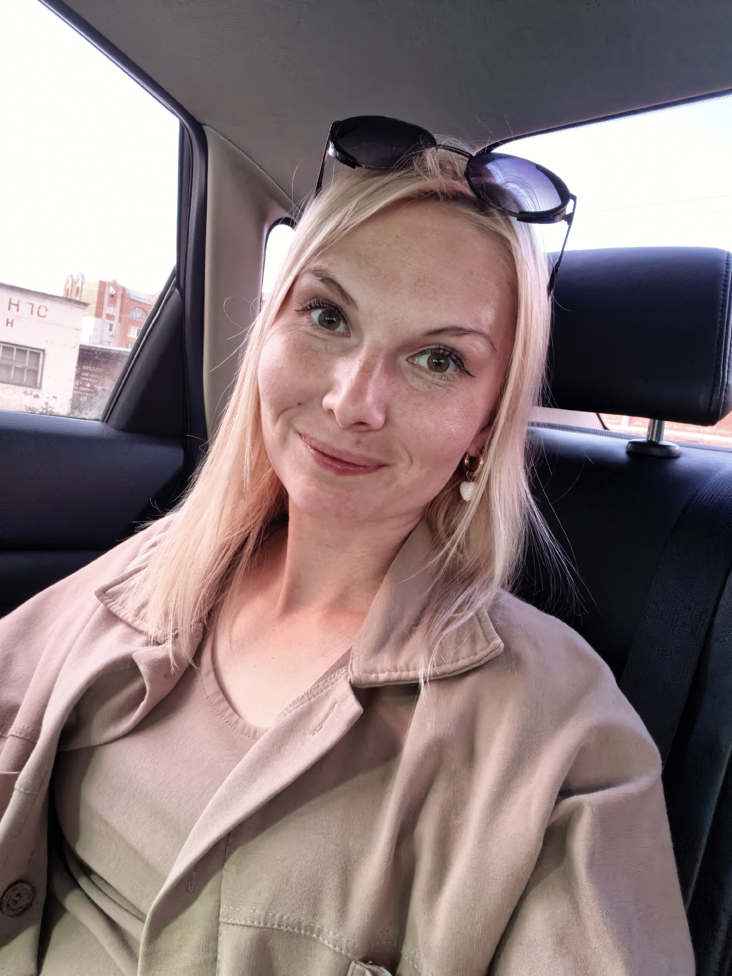 An outdoor, eye-level, medium shot captures a blonde woman in her late 20s to early 30s, seated in the back of a car. She is looking towards the camera with a slight smile, her head tilted slightly to her right. Her light brown eyes are open, and her eyebrows are well-defined. Her blonde hair, styled straight, falls around her shoulders. Sunglasses are perched on top of her head. She is wearing a light beige button-up jacket over a matching t-shirt. A small, heart-shaped white earring is visible on her left ear.
The car interior features a dark gray headrest behind her. Through the window to her left, a building with white and red brickwork is visible, along with a hint of a streetlight. The building has windows and what appears to be Cyrillic lettering on its facade. The scene suggests a daytime setting. The provided location is Sterlitamak, Russia.
