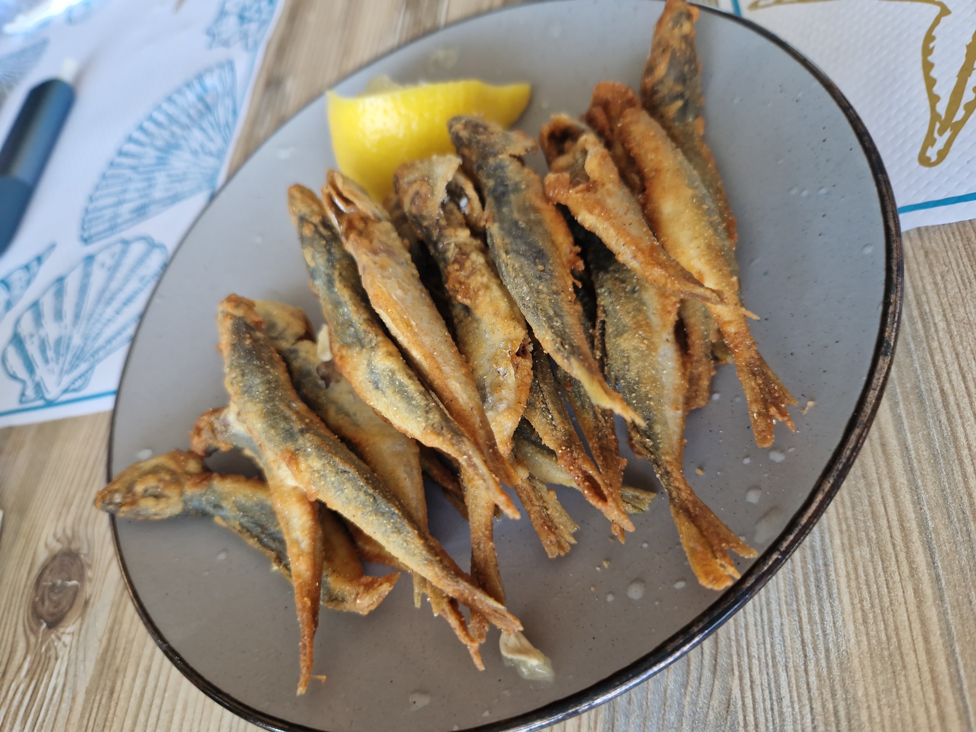 A plate of fried fish is displayed on a wooden table. The fish are small, whole, and appear to be coated in a light batter or seasoned flour. They are arranged in a pile on a gray plate with a dark rim. A wedge of yellow lemon is placed on the right side of the plate. The table surface has a visible wood grain pattern. In the background, a white napkin with blue seashell designs is visible, as well as a blurred blue object. The scene is likely from a restaurant in Pomorie, Bulgaria, given the proximity to the Black Sea, a region known for fresh seafood.