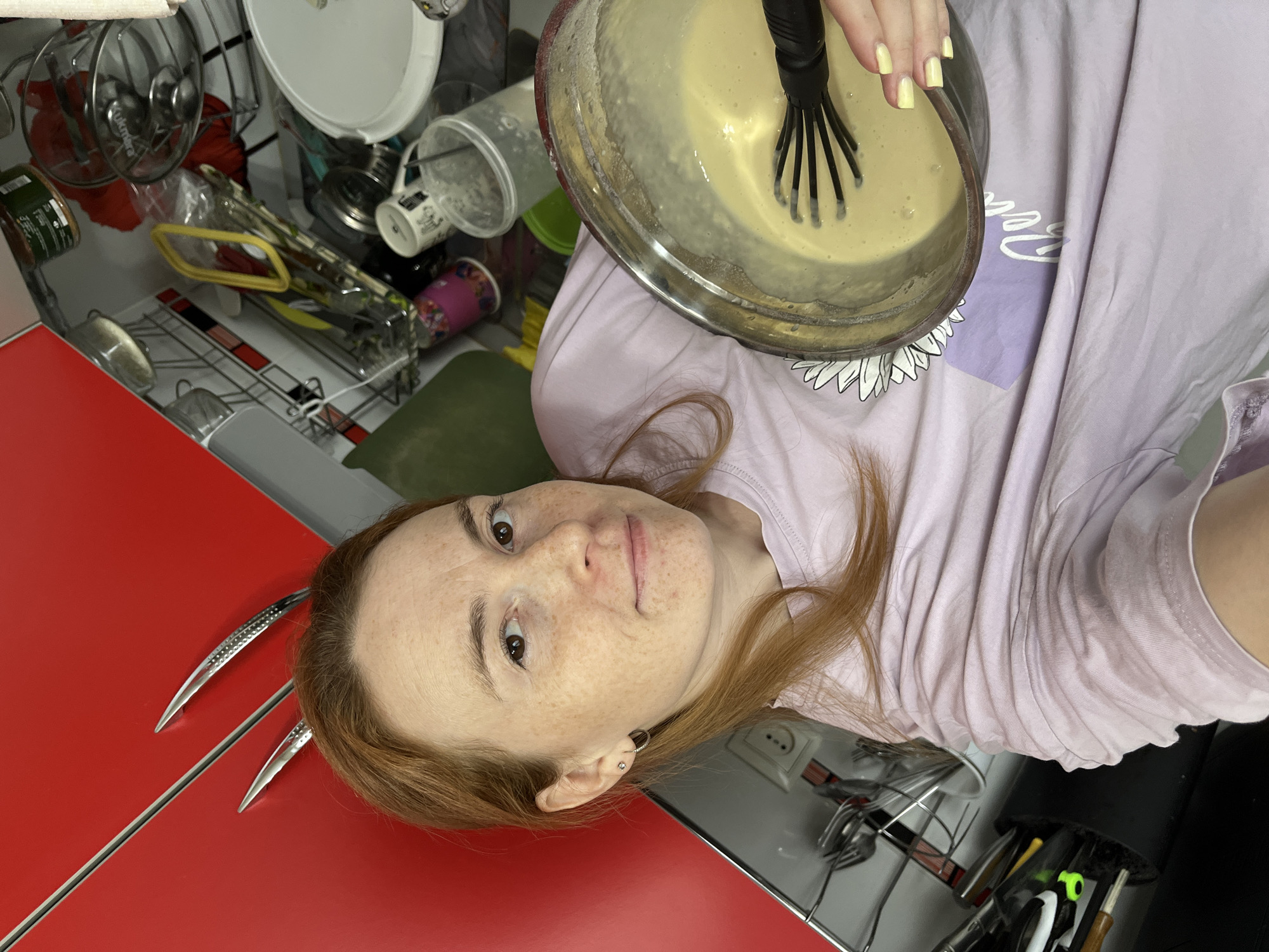 In a residential kitchen in Chaikovsky, Russia, a female adult subject with reddish-brown hair, fair skin, and freckles is depicted. She wears a light lavender short-sleeved t-shirt featuring a partially visible white graphic. Her fingernails are painted a pale yellow. The subject is holding a clear glass mixing bowl containing a light yellow, semi-viscous liquid, with a black whisk submerged within it. She is looking directly towards the camera, suggesting a self-portrait. The background on the left consists of a reflective red cabinet or wall panel with three chrome-colored, elongated decorative elements. To the right, a cluttered kitchen counter is visible, containing a white European-style electrical outlet, a utensil holder, glass and plastic containers, a dish drying rack with assorted glassware, and several mugs.