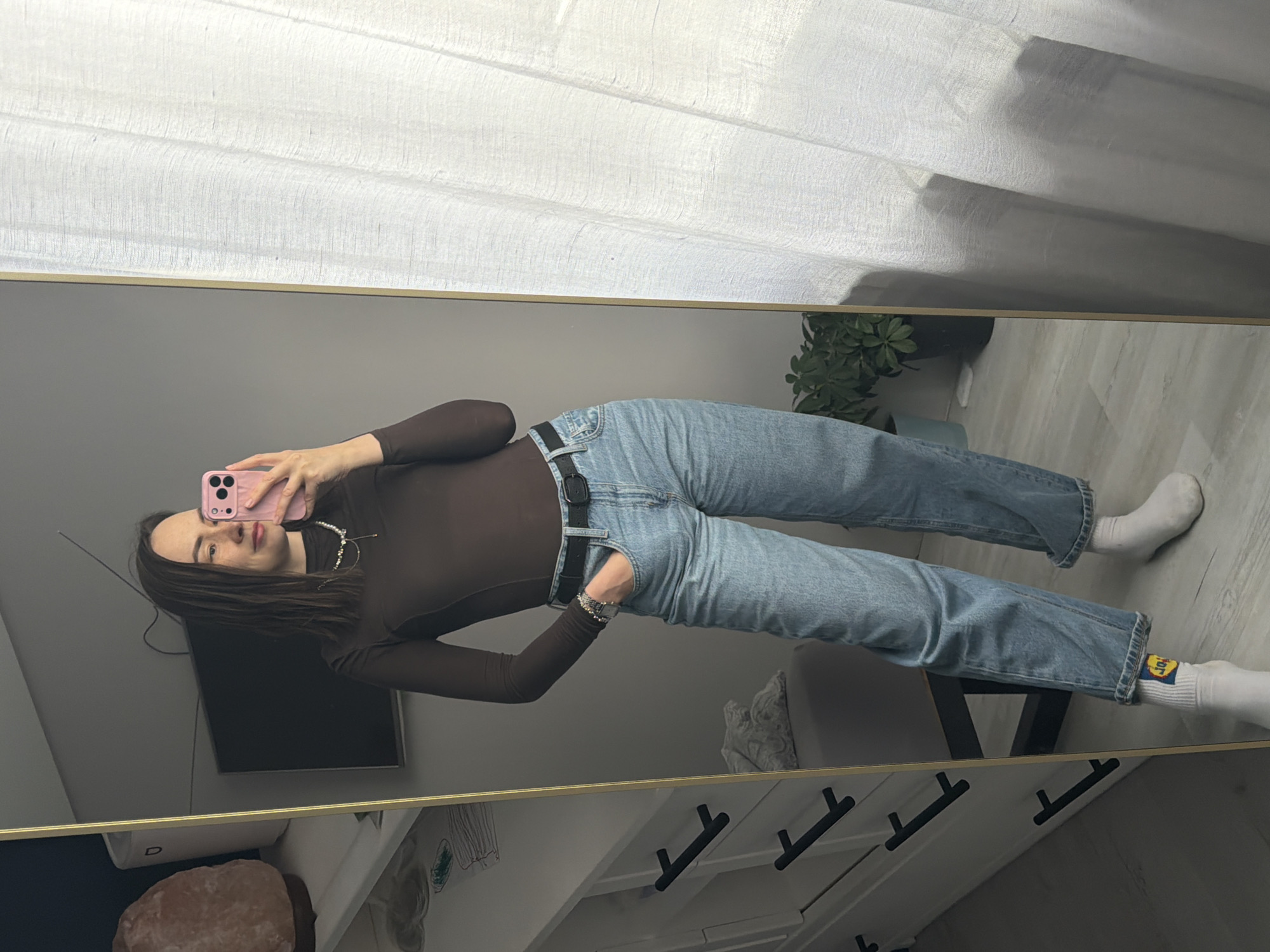 A young woman is depicted taking a mirror selfie indoors, appearing to be in Chaikovsky, Russia. She stands full-length, holding a pink smartphone to capture her reflection. She is dressed in a dark brown long-sleeved top, light blue jeans, and white socks, with a black belt securing her jeans. A silver watch is visible on her left wrist, and she wears a delicate, layered necklace.
Her reflection shows her with dark hair, a subtle smile, and her eyes looking towards the phone. The background, also a reflection, reveals a bright room with white sheer curtains, suggesting a window and natural light. Other reflected objects include a potted green plant, a black flat-screen television, a white cabinet with black drawer handles, and a large pink/orange crystal lamp.
A distinctive detail is the logo visible on her white socks, featuring a yellow circle with a blue square inside, bearing the white text "LIDL." The overall impression is a casual, well-lit indoor setting, likely a home, with the woman presenting her outfit.