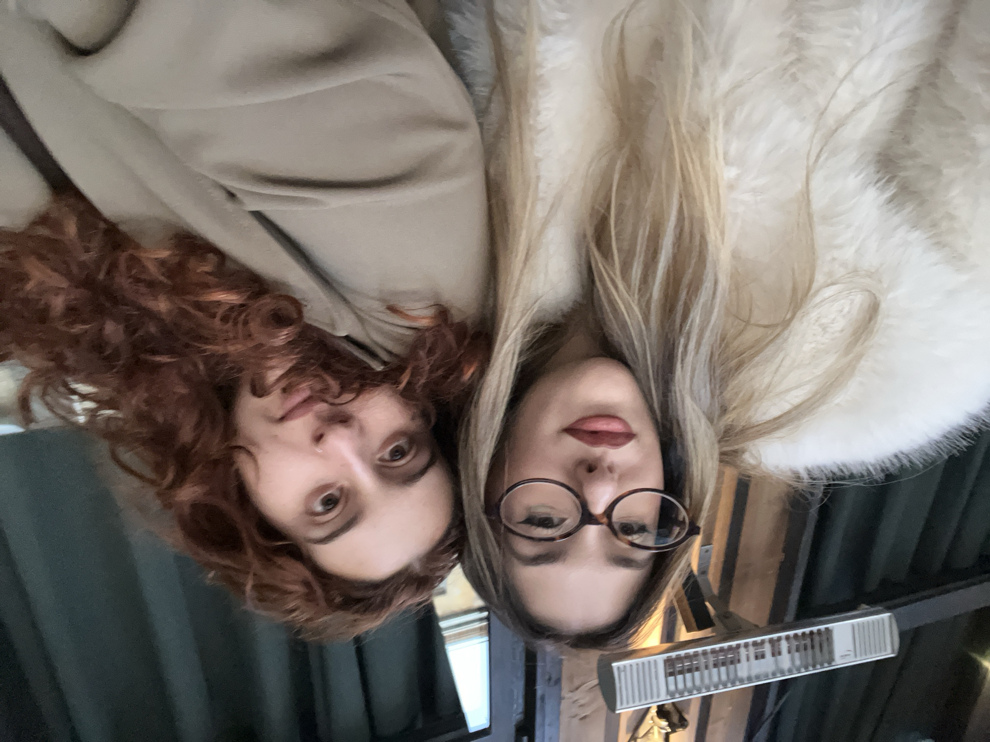 The image shows two young women posing for a close-up photo, looking directly downwards at the camera. The setting appears to be indoors, possibly a cafe or restaurant, given the presence of a heater and wooden architectural elements in the background.
On the left, a woman with voluminous, reddish-brown curly hair is visible. She is wearing a light brown or tan-colored garment. Her gaze is directed at the viewer, with a neutral to slightly pensive expression.
Next to her, on the right, is another woman with long, straight blonde hair. She is wearing distinctive round, tortoise-shell rimmed glasses. Her lips are slightly parted, revealing red lipstick, and she has a relaxed facial expression. She is dressed in a fluffy, white faux fur jacket or coat.
The background behind them is softly blurred, showing dark, possibly wooden beams and an indistinct ceiling or wall. A modern, silver-toned rectangular heater is mounted on a dark structure in the lower right portion of the frame. The overall mood is casual and intimate. No visible text is discernible in the image. The location context is Sofia, Bulgaria.
