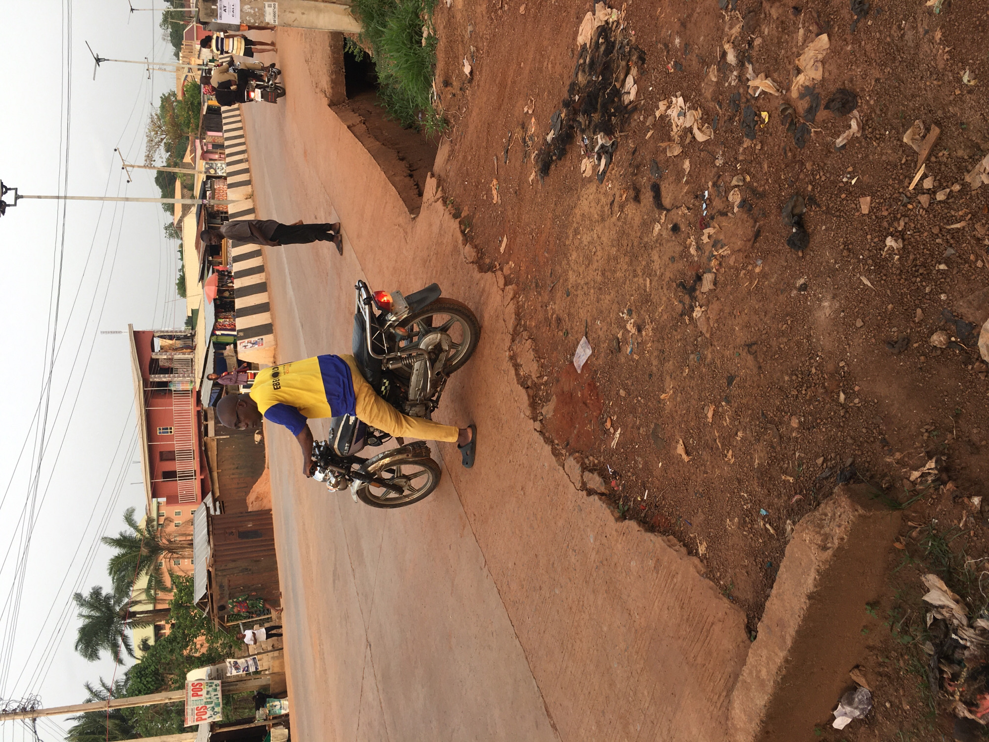 This image captures a daytime roadside scene in Isieke, Nigeria, likely in the late afternoon under an overcast sky. The photograph is oriented vertically.
In the foreground, a man wearing a yellow t-shirt with blue sleeves and yellow pants is riding a black motorcycle on a light-colored paved road. His t-shirt has partial text "EBRO..." visible. Further down the road, another man in dark attire stands near the roadside, and additional figures are discernible in the distance near buildings.
To the right of the paved road, there is a wide dirt shoulder and an embankment that is heavily littered with various types of rubbish, including plastic bags, paper, and organic debris. A concrete drainage channel runs parallel to the road.
The background reveals a mix of buildings, including a noticeable red multi-story structure, and smaller roadside stalls, some with corrugated metal roofs. Utility poles with extensive power lines crisscross the upper portion of the frame. Palm trees and other greenery are scattered throughout the setting.
Visible text includes "POS" on a white sign attached to a pole in the lower left, and "AT CALL" along with a partial phone number "08144000140" on a sign attached to a utility pole further down the road. The scene portrays a typical daily street activity in a bustling local environment.