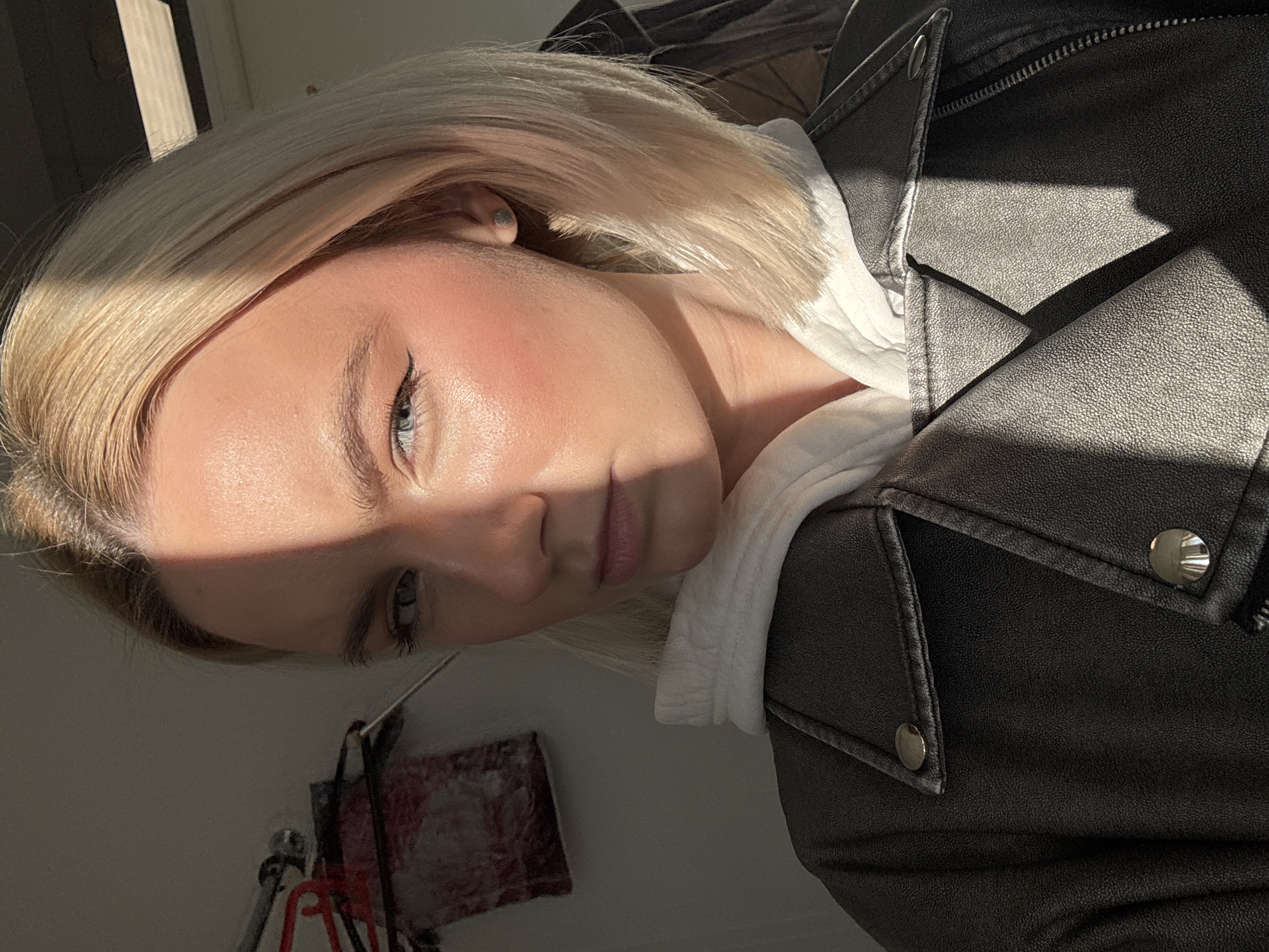 A portrait of a person with blonde hair and light skin, wearing a white hoodie under a black leather jacket, is partially illuminated by sunlight. The background includes an indistinct wall and a small, blurred red and white object. The scene does not provide enough detail to suggest a location in St. Petersburg, Russia.
