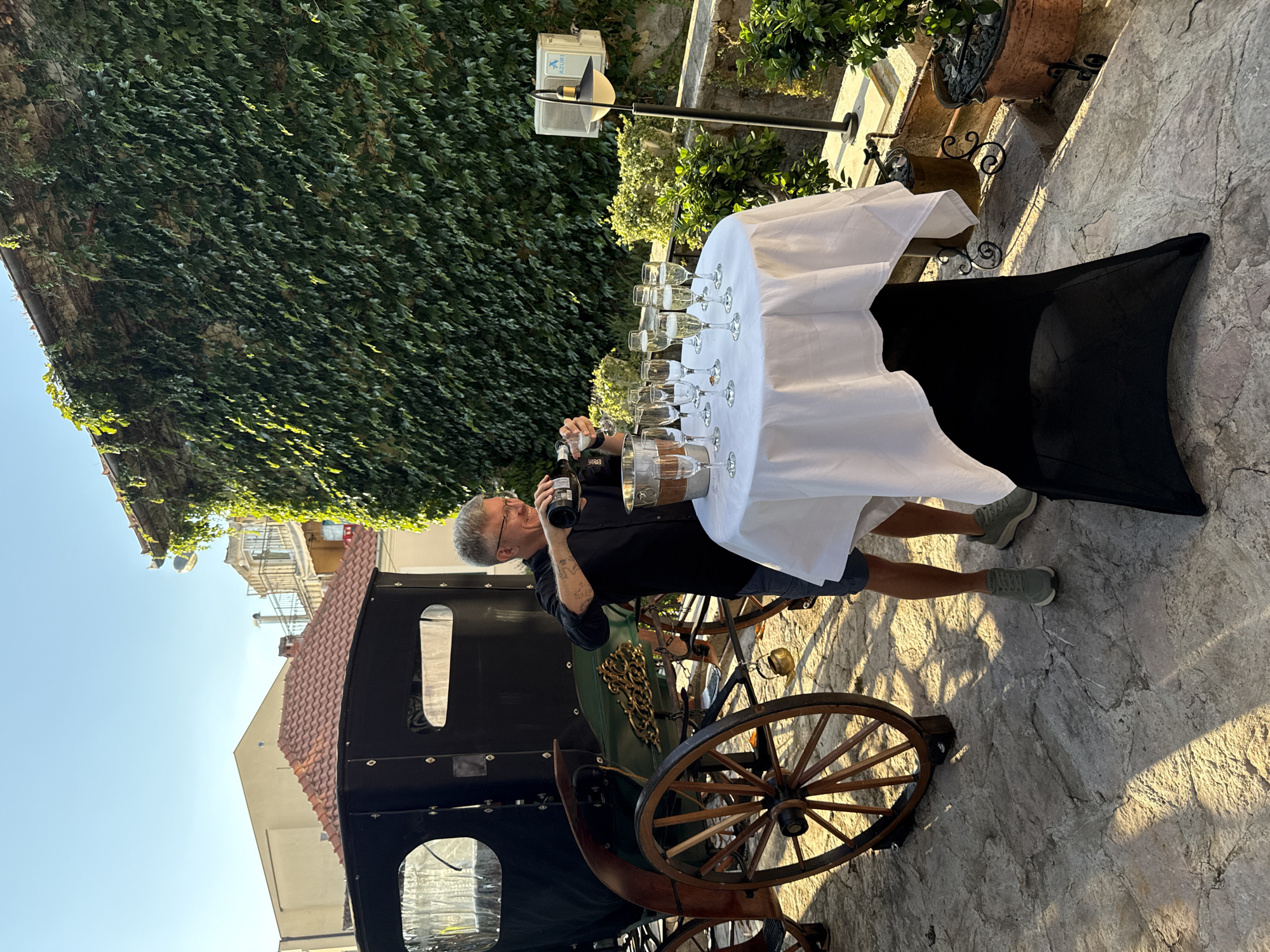 In Herceg Novi, Montenegro, a man in a black shirt and shorts is pouring champagne from a bottle into a flute glass. He is standing beside a white-covered round table, which has an ice bucket and several other flute glasses filled with champagne on it. Behind him, a dark green horse-drawn carriage is parked. The scene takes place outdoors, with a stone pathway, lush green ivy covering a wall, and buildings visible in the background. The lighting suggests it is late afternoon or early evening.