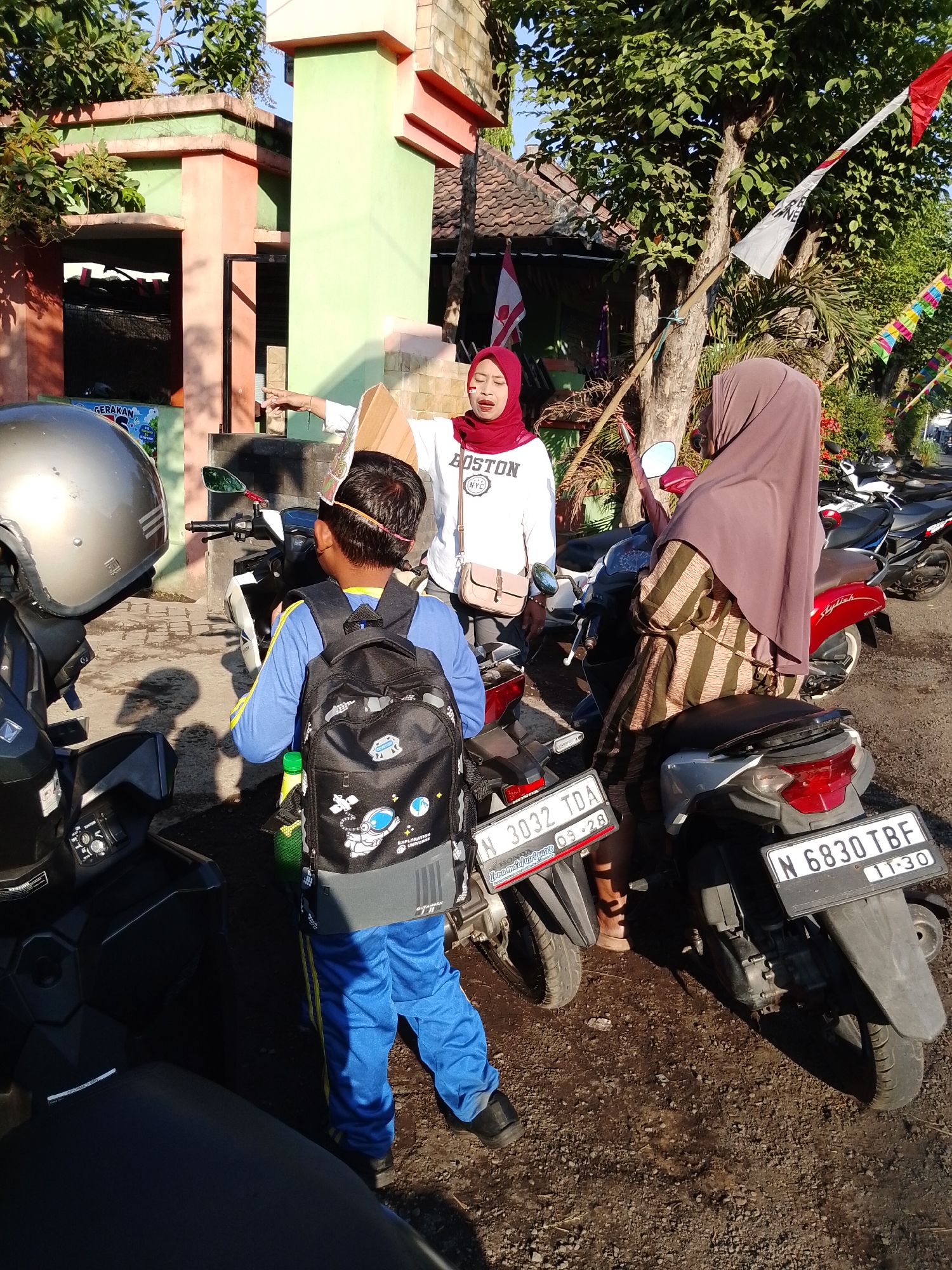In Pasuruan, Indonesia, on a sunny day, a young boy wearing a blue long-sleeved shirt, blue pants with yellow stripes, and a black backpack with a space-themed design stands next to a motorcycle. He is wearing glasses and black shoes. To his right, a woman in a maroon hijab and a striped dress is sitting on another motorcycle, facing away from the camera. Another woman in a red hijab and a white "BOSTON NYC" sweatshirt stands between them, holding a cardboard cutout. The background features a building with green and orange accents, trees, and flags. Several other motorcycles are parked in the vicinity.