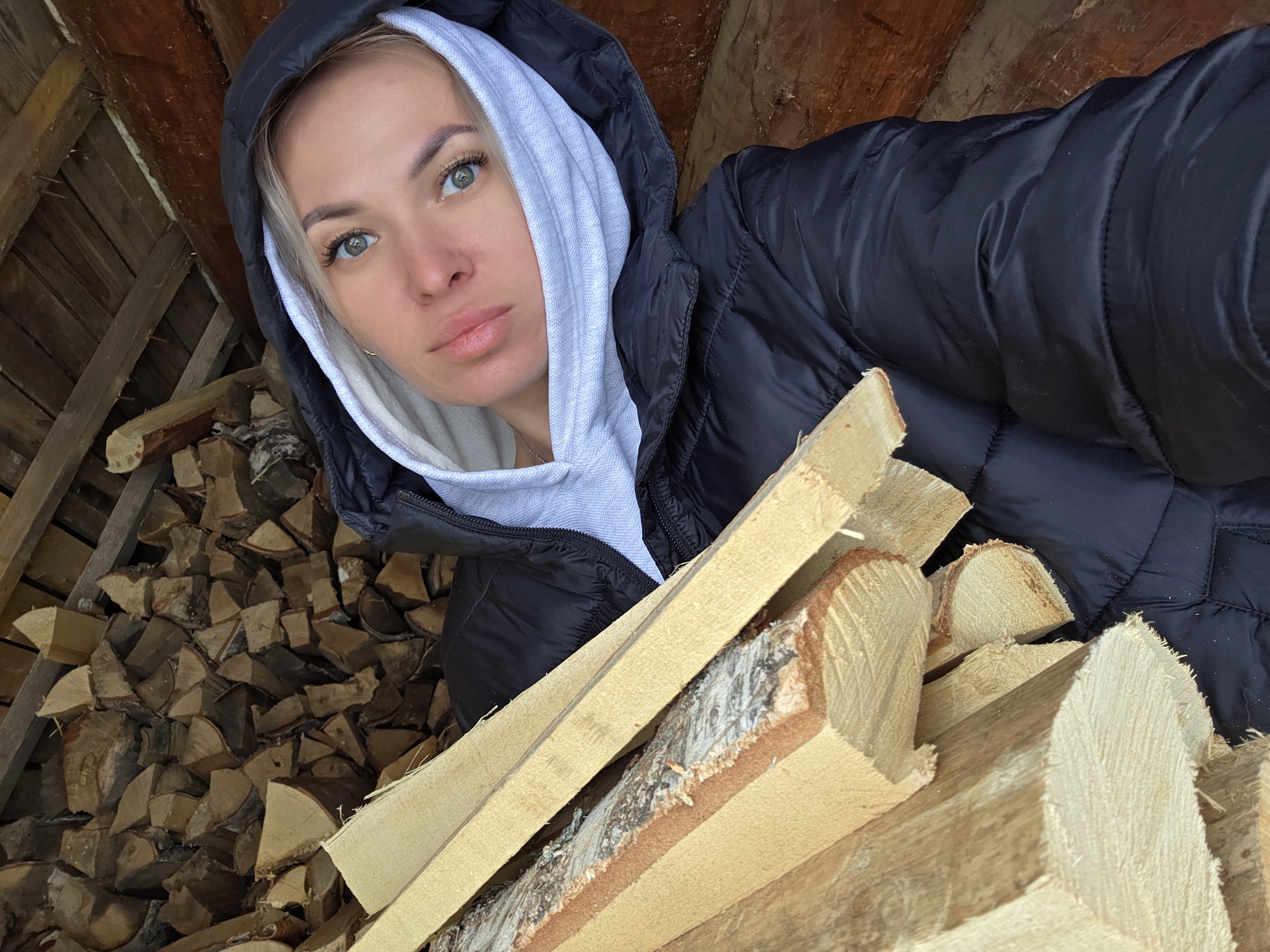 The image shows a young woman in a black puffer jacket with a gray hooded sweatshirt underneath. She is looking at the camera with a neutral expression. The background shows stacked firewood, both behind her and held in front of her, suggesting she is in or near a wood shed or storage area. The presence of cut wood and the style of construction visible in the background indicate a potentially rural or semi-rural location. Given the prompt specifies Chaikovsky, Russia, it is likely this image was taken in a location near or within that city with access to firewood for heating purposes.