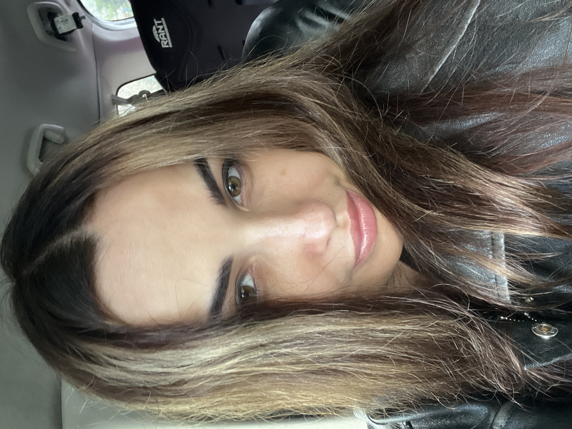 A young woman is prominently featured in a close-up portrait, taken inside what appears to be a vehicle. Her head is tilted, and she looks directly at the camera with a subtle, closed-mouth smile. She has long, dark brown hair with lighter, possibly balayage or highlighted strands, particularly visible around her face and ends. Her eyebrows are dark and well-defined, complementing her brown eyes. She has clear skin and is wearing light pink lip gloss or lipstick.
She is dressed in a dark, textured jacket, which appears to be leather, with a small metallic charm or zipper detail visible near her chin. The background shows parts of a car interior, including a light-colored ceiling and a grab handle. Behind her, a black headrest or car seat is visible, bearing the white text "RANT." The lighting suggests daylight, entering from unseen windows. The overall impression is a casual selfie or portrait taken while seated in a car.