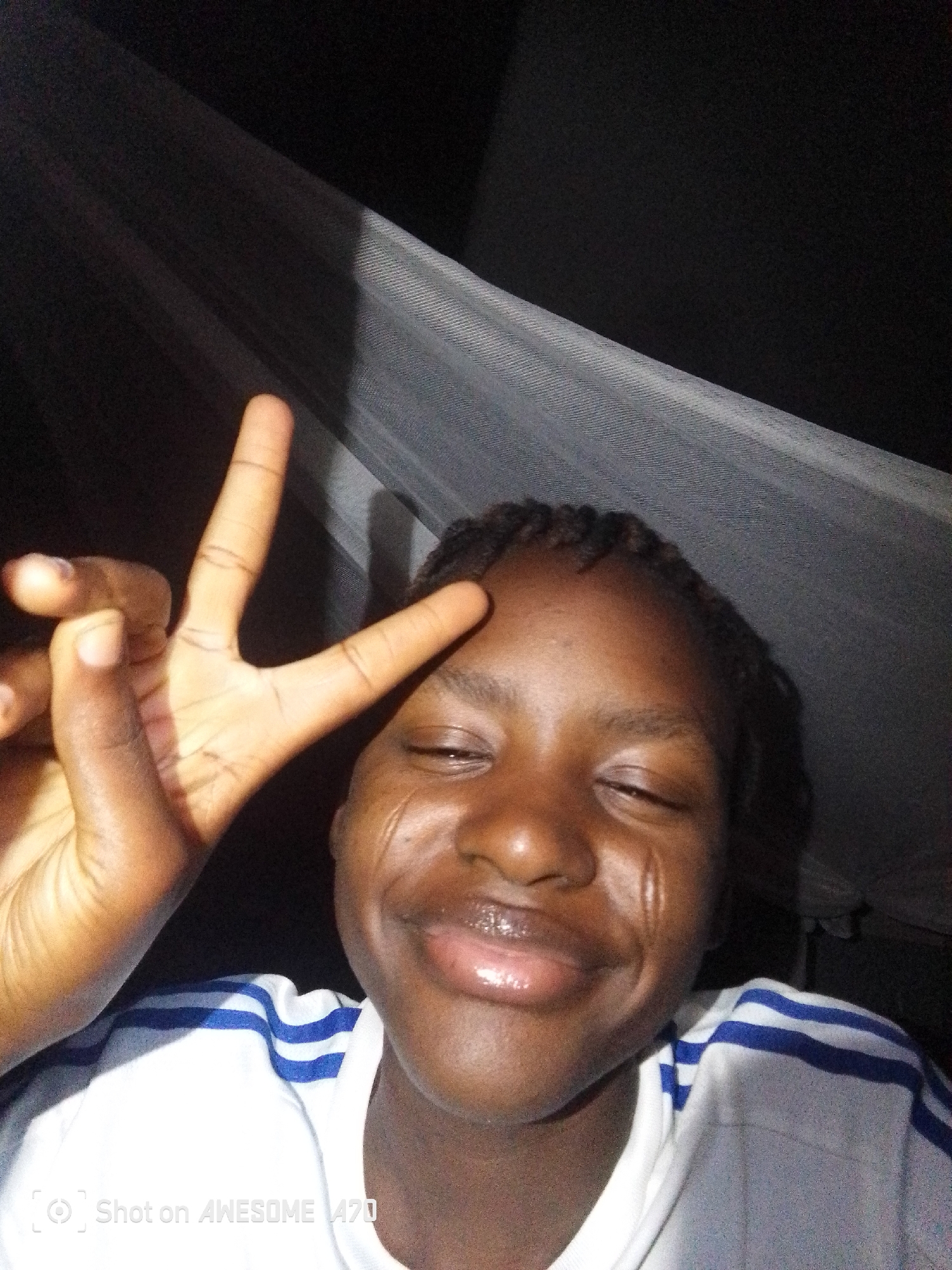 A close-up shot depicts a person with dark skin, potentially a teenager or young adult, positioned indoors under a light-colored, translucent fabric that might be a mosquito net. The person's hair is braided, and they are wearing a white T-shirt with blue stripes on the shoulders. They are making a "peace" or "victory" sign with their left hand, held up near their forehead. The subject is smiling and appears to be facing the camera directly. The background is predominantly dark, with the lighter fabric being the primary source of illumination in the scene. At the bottom of the image, there is a translucent "Shot on AWESOME A70" overlay. The setting is in the city of Unknown, Unknown.