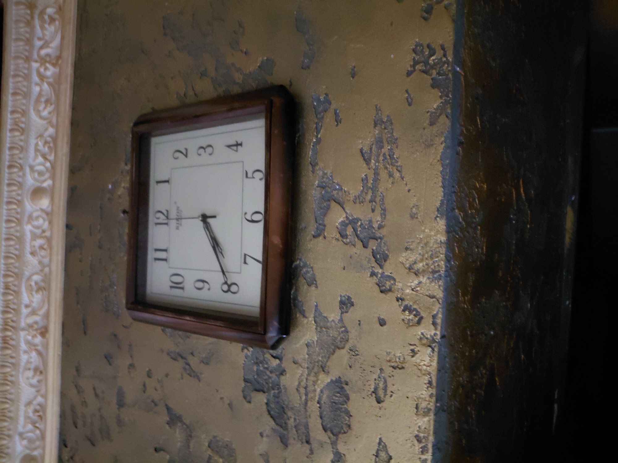 The image displays a close-up, low-angle shot of a square-faced clock mounted on a textured, gold-colored wall. The wall exhibits a distressed appearance with peeling paint or plaster, revealing darker areas underneath. Above the clock, a decorative, ornate molding with intricate detailing is visible. The clock itself has a dark wooden frame and a white face with black Arabic numerals. The hour hand points to approximately 3:40, and the minute hand is close to the 8. The background is dark and out of focus, suggesting the clock is indoors. There are no people or animals present in the image, and no specific location information is discernible from the visual content.