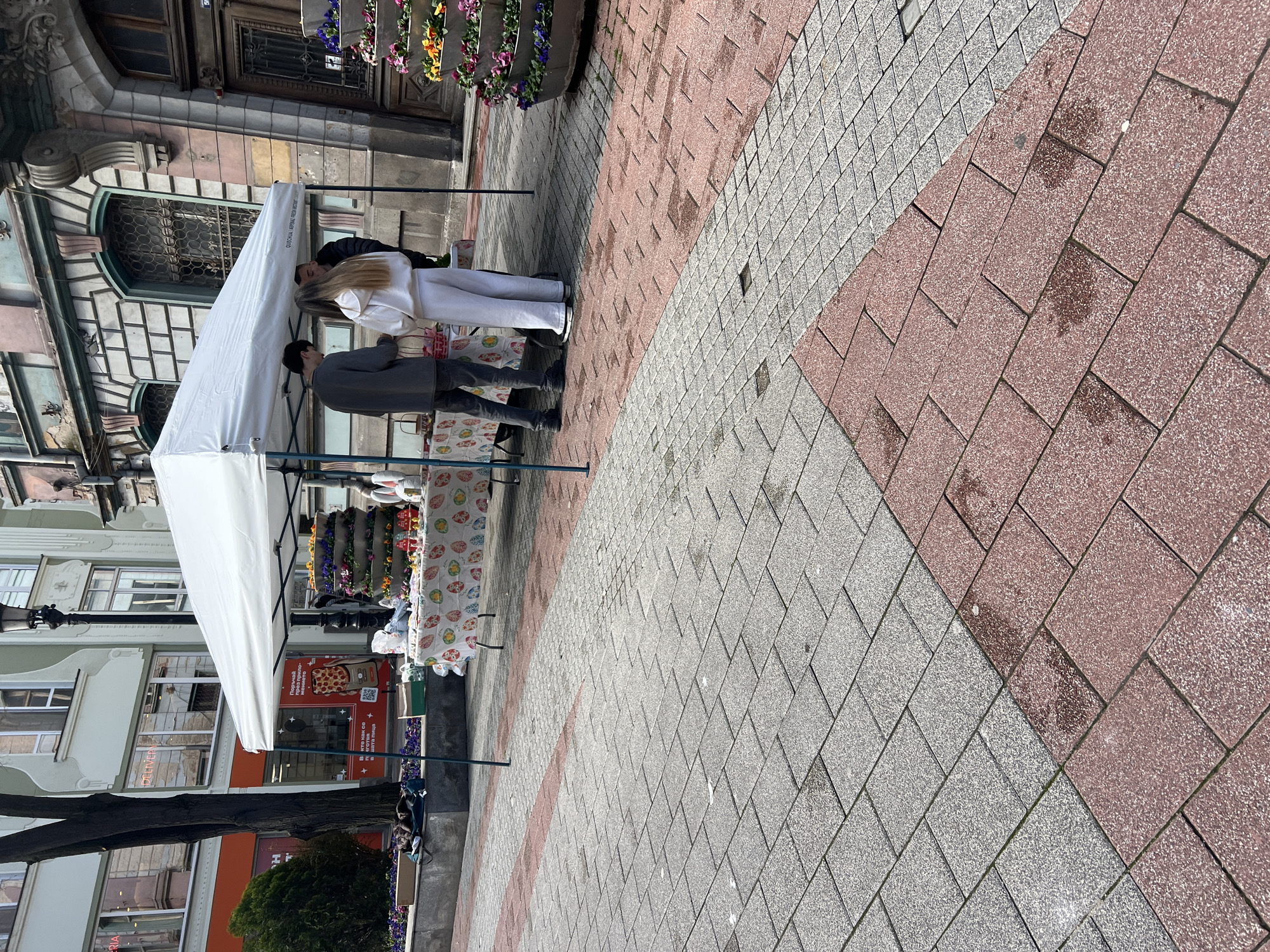 The image captures a street scene in Plovdiv, Bulgaria. A white tent is set up on a paved area, possibly for a small market or sale. Two people are seen standing near the tent. One is dressed in a grey jacket and dark trousers, and the other in a white outfit. The individuals appear to be engaged in a transaction or conversation related to the display on the table under the tent, which has a patterned tablecloth. The background includes buildings with detailed architectural elements, flower planters, and advertisements for a pizza restaurant. The pavement is made of alternating sections of red and gray stones.