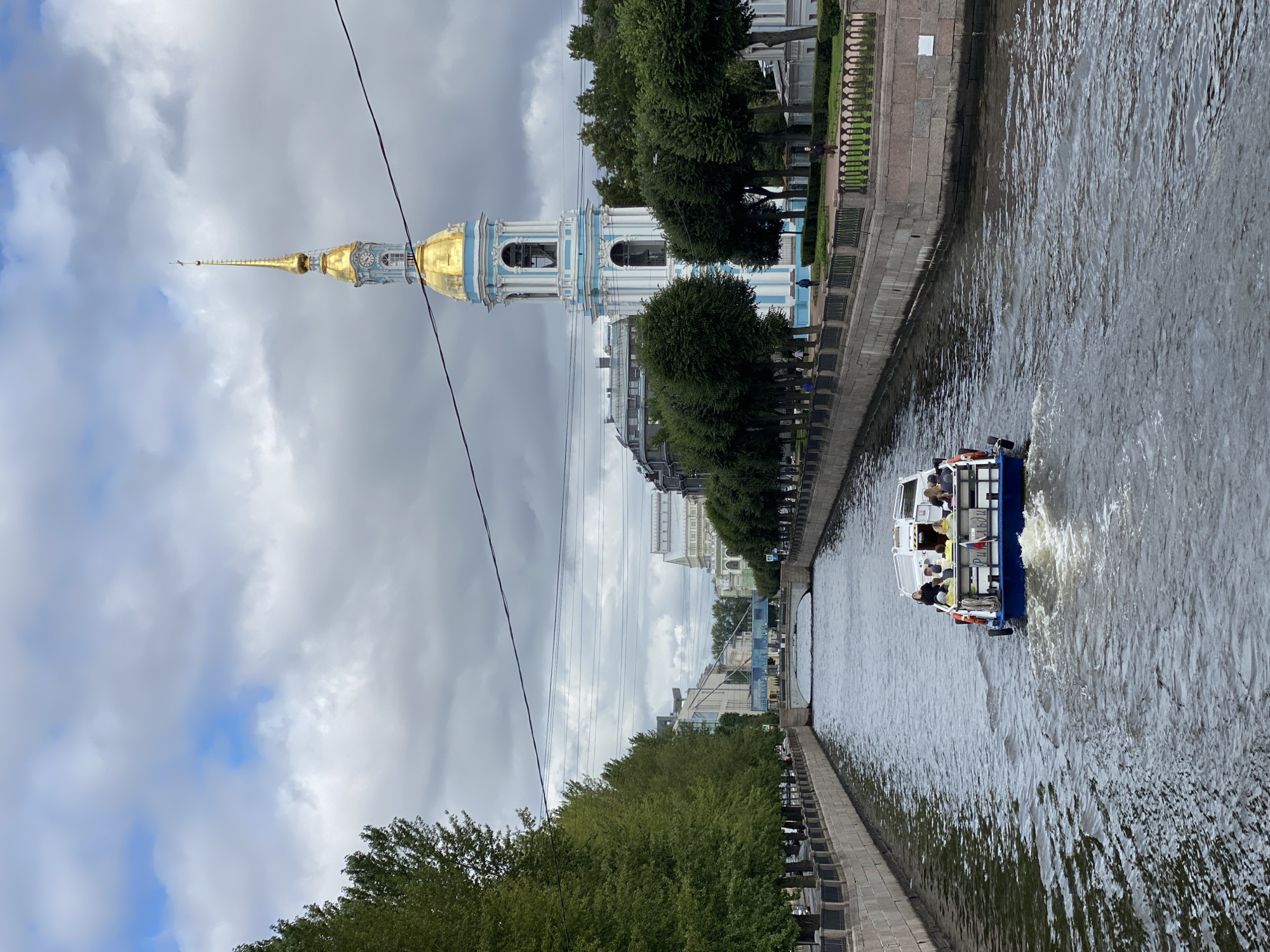 This image captures a view of the Griboyedov Canal in Saint Petersburg, Russia, with the iconic St. Nicholas Naval Cathedral's bell tower dominating the background. A tour boat, carrying several passengers, moves away from the viewer down the canal. The water is choppy, indicating the boat's movement. The canal banks are lined with stone walls and manicured trees. In the distance, a bridge spans the canal. The sky is overcast with patches of blue peeking through, suggesting a cloudy day. The architecture of the buildings lining the canal is characteristic of Saint Petersburg, with a mix of historical and more modern structures.