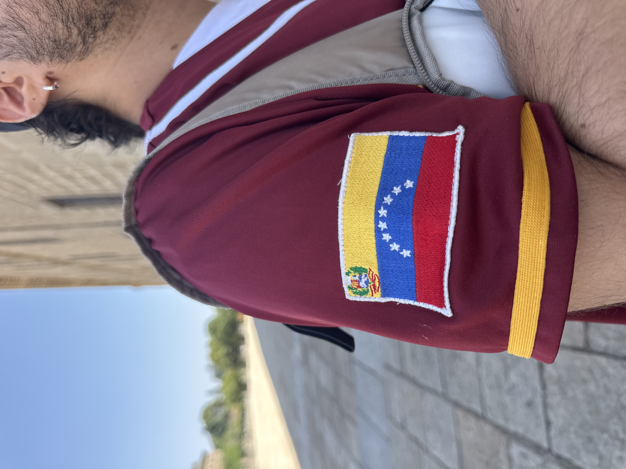 A man is shown from the chest down, wearing a maroon shirt with a Venezuelan flag patch on the sleeve. He has dark hair and a beard and is wearing a silver hoop earring. He is standing in a courtyard with a paved ground and a stone building in the background. The sky is blue. The image appears to be taken in the city of San Lorenzo de El Escorial, Spain, likely within the grounds of the Royal Site of San Lorenzo de El Escorial.
