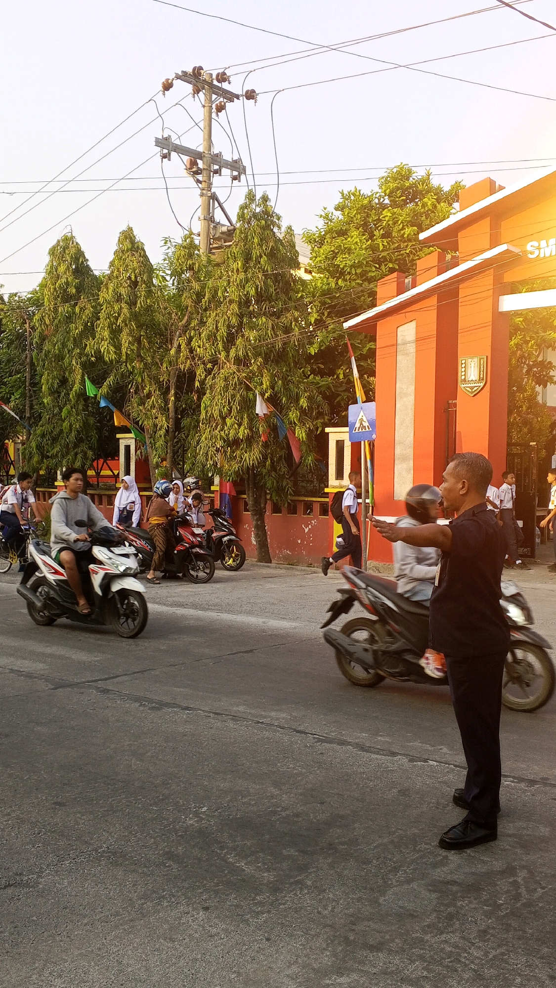 This image depicts a street scene in Semarang, Indonesia, likely near a school entrance. The foreground shows a man in a dark blue shirt and dark pants, wearing black shoes, gesturing with his right arm extended, likely directing traffic. He is standing on the right side of the road, facing left. To his left, a motorcycle rider wearing a gray hoodie and shorts is on a white scooter, heading towards the left of the frame. Behind this scooter, a group of students in school uniforms are on motorcycles. Some are wearing white shirts and dark pants or skirts, while others wear hijabs. They are congregated near an orange building with white trim, which appears to be an institutional entrance with decorative flags. Trees line the area behind the school building. In the background, a utility pole with numerous wires stretches across the sky, and power lines are visible. The road surface is asphalt with visible cracks and wear. The overall lighting suggests it is daytime, possibly late afternoon, with a warm glow due to sunlight.