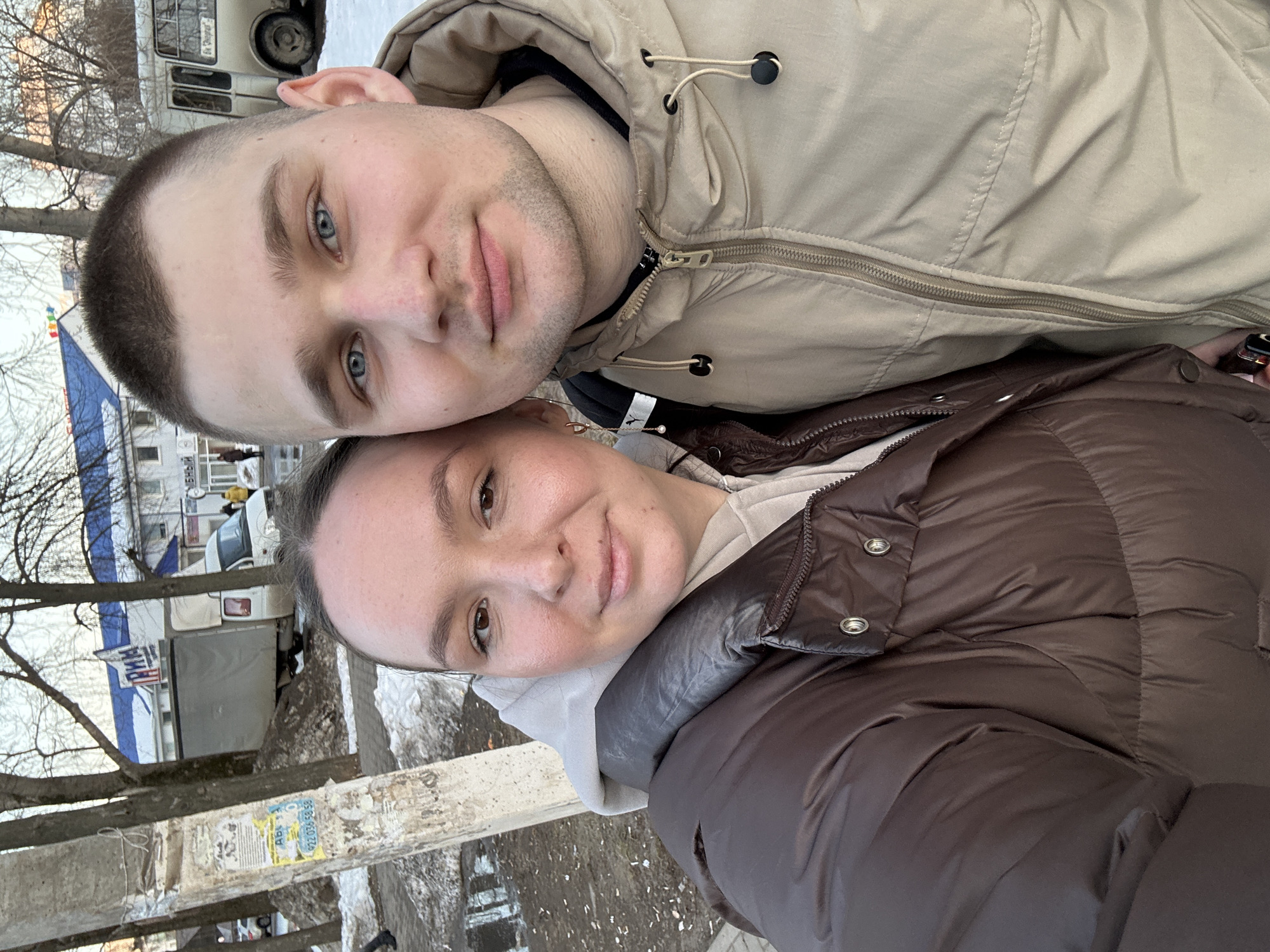 A selfie-style portrait shows a male and female subject outdoors in an urban setting in Chaikovsky, Russia, during what appears to be late winter or early spring. The female, positioned on the left and slightly closer to the camera, wears a brown puffer jacket over a light grey hooded sweatshirt. She has brown eyes and light skin, with her hair pulled back. The male, on the right, wears a tan hooded jacket over a dark collar. He has short brown hair, blue eyes, and light stubble. Both subjects look directly at the camera.
The background features a concrete utility pole on the left, adorned with various faded papers, one displaying the phone number "922 036 58 58." The ground consists of a sidewalk and dirt patches with melting snow and puddles. Vehicles visible include a white cargo van with a grey canvas top behind the pole, and a white passenger bus bearing the Cyrillic text "ВК "Опора"" on its side, partially obscured on the far right. A two-story white building with a blue roof is prominent in the background, featuring a large blue sign with the Cyrillic word "ПРИМА" (PRIMA) at its top, indicating its likely location on Mira Street 42. Bare deciduous trees are scattered throughout the scene under an overcast sky.