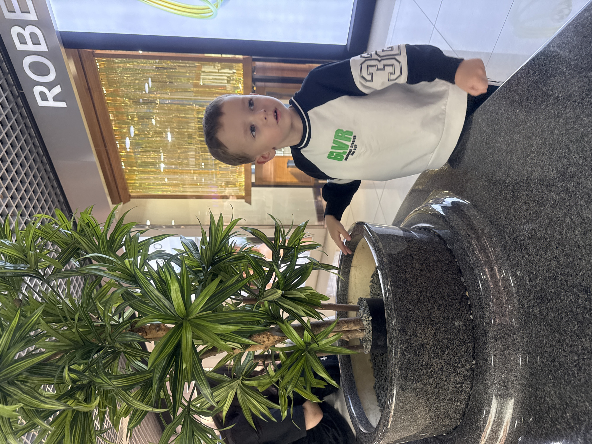 The image captures an indoor scene, likely within the Clover House shopping center in Vladivostok, Russia. A young male child, approximately three to five years old, stands beside a large artificial plant with long, variegated green leaves. The plant is housed in a multi-tiered circular planter constructed from dark gray speckled granite, with fine gray pebbles surrounding the plant's base. The child wears a white and dark blue long-sleeved raglan sweatshirt, featuring green text "GLVR" on the chest and a white outlined number "33" on the right sleeve. His right hand rests lightly on the planter's edge as he looks towards the upper right of the frame. In the background, a store sign partially displays "ROBE" in white letters against a dark background, positioned above a shimmering, golden-yellow vertical strip display. A person wearing dark clothing is partially visible behind the plant on the left. The floor is composed of light-colored, reflective tiles, and the ceiling features a dark grid pattern with integrated lighting.