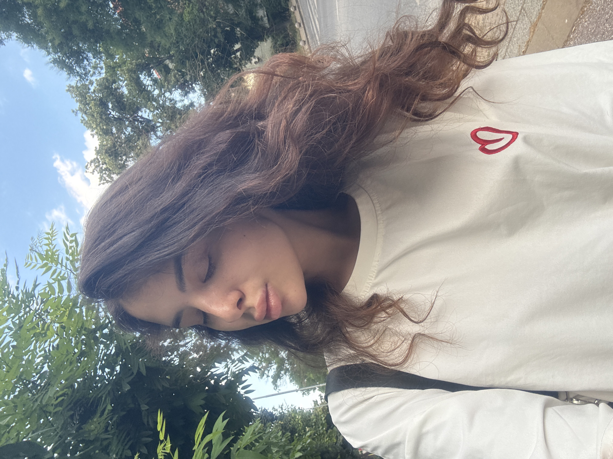 A young woman with long, wavy brown hair is taking a selfie outdoors. She is wearing a white t-shirt with a small red heart emblem on the left side of her chest. Her eyes are closed, and her head is tilted slightly downwards. In the background, lush green trees and a blue sky with scattered clouds are visible, suggesting an urban park or a tree-lined street. The context of Plovdiv, Bulgaria is not evident from the visual information in the image.