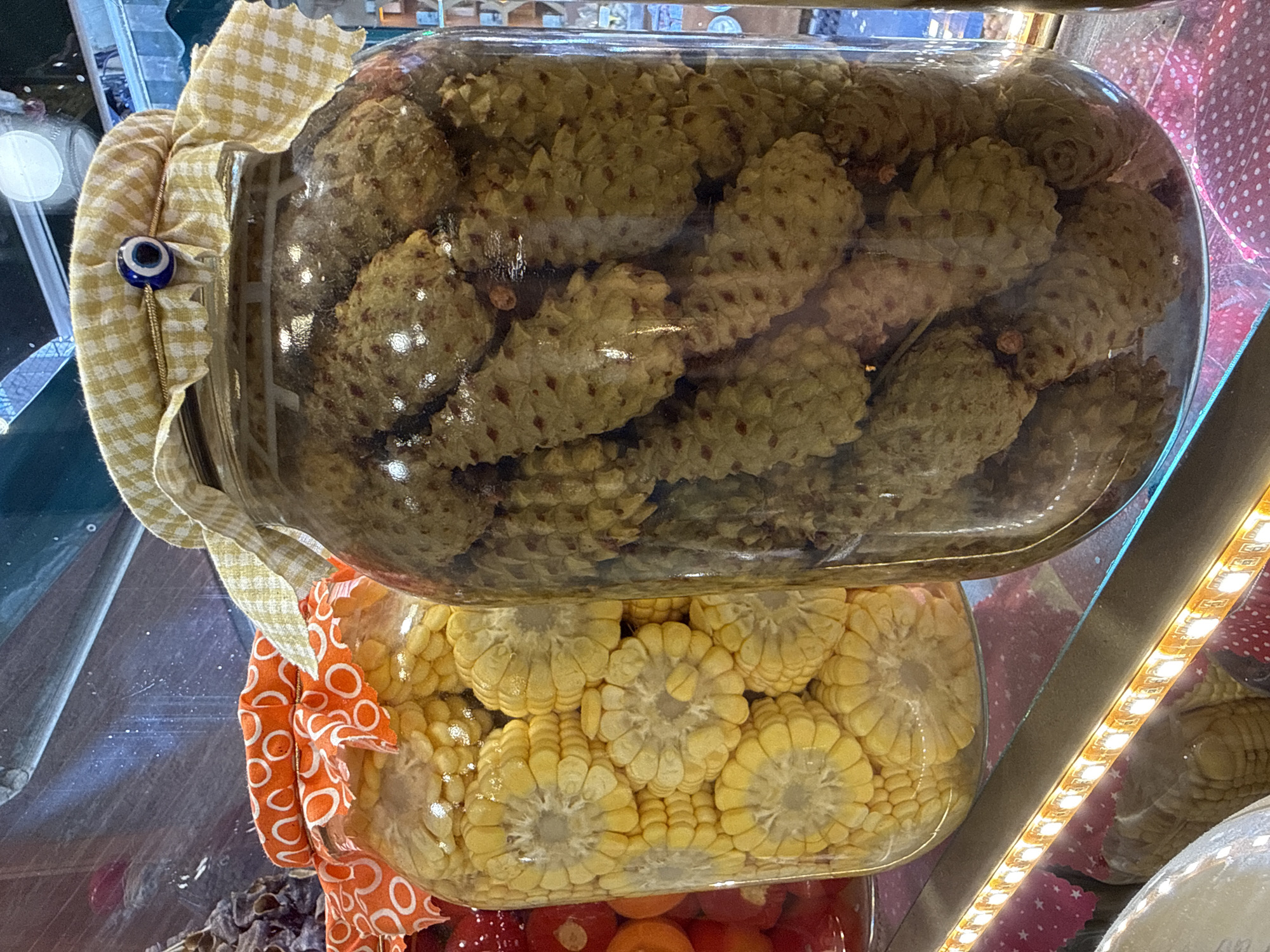 Two large glass jars are visible in the foreground of this medium shot, with the jar on the right being more prominent. The jar on the right is filled with what appear to be pickled pine cones, submerged in a clear liquid. The pine cones are light brown and have a textured, layered appearance. The jar is sealed with a yellow and white checkered cloth, tied with twine, and adorned with a blue and white evil eye charm. The jar on the left contains slices of corn on the cob, also submerged in liquid. These corn slices are a bright yellow and arranged vertically within the jar. This jar is covered with an orange and white patterned fabric, secured similarly with twine.
Behind these jars, other items are partially visible, including what appears to be a jar of red peppers to the left of the corn, and a dark, lumpy item in a jar behind the pine cones on the far left. The background is indistinct, but suggests a display or market setting, with shelves and possibly other food items. A strip of LED lights runs horizontally across the bottom of the image, illuminating the display. The overall scene is brightly lit, with the glass jars reflecting the surrounding light.
This image was taken at a spice shop in Kadıköy, Istanbul, Turkey, located on the Asian side of the city. Kadıköy is a bustling district known for its vibrant markets, restaurants, and cultural scene. The presence of pickled vegetables and other preserved goods is common in such establishments in Turkey.