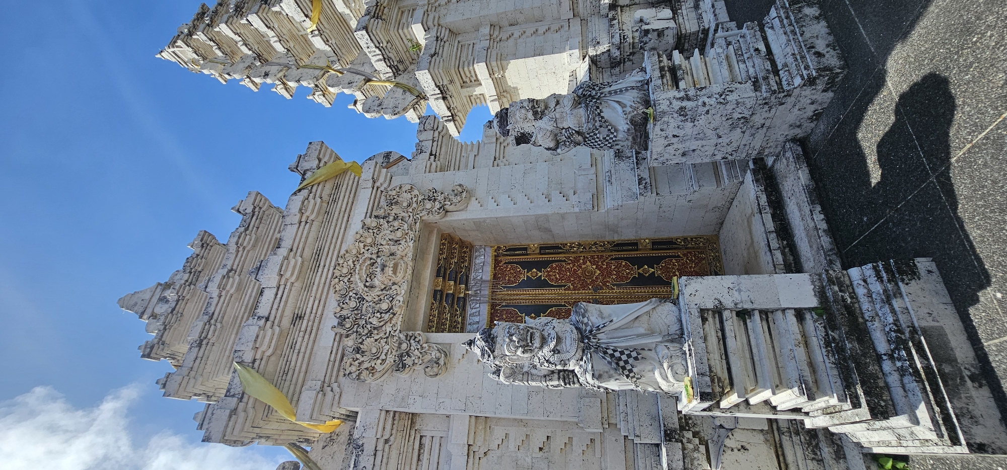 A low-angle shot shows the entrance to a Balinese temple in Kuta Selatan, Indonesia. The temple is constructed of white stone with intricate carvings. Two statues of deities flank the entrance, wearing checkered cloths. A pair of ornately decorated wooden doors are visible inside the entrance. Yellow cloths are draped across the temple's facade. The sky above is clear and blue, with the edge of a shadow partially visible in the top-right corner of the frame.