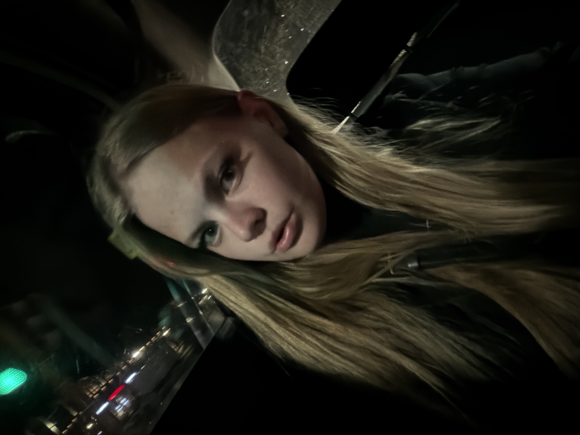 A young female with long blonde hair is looking towards the camera. She is wearing a dark shirt. Her face is lit from the front, and her eyes are green. The background is dark and blurred, with indications of car lights and a traffic light visible through a window. This scene appears to be inside a vehicle at night. The location is identified as Pervomaysky, Russia.