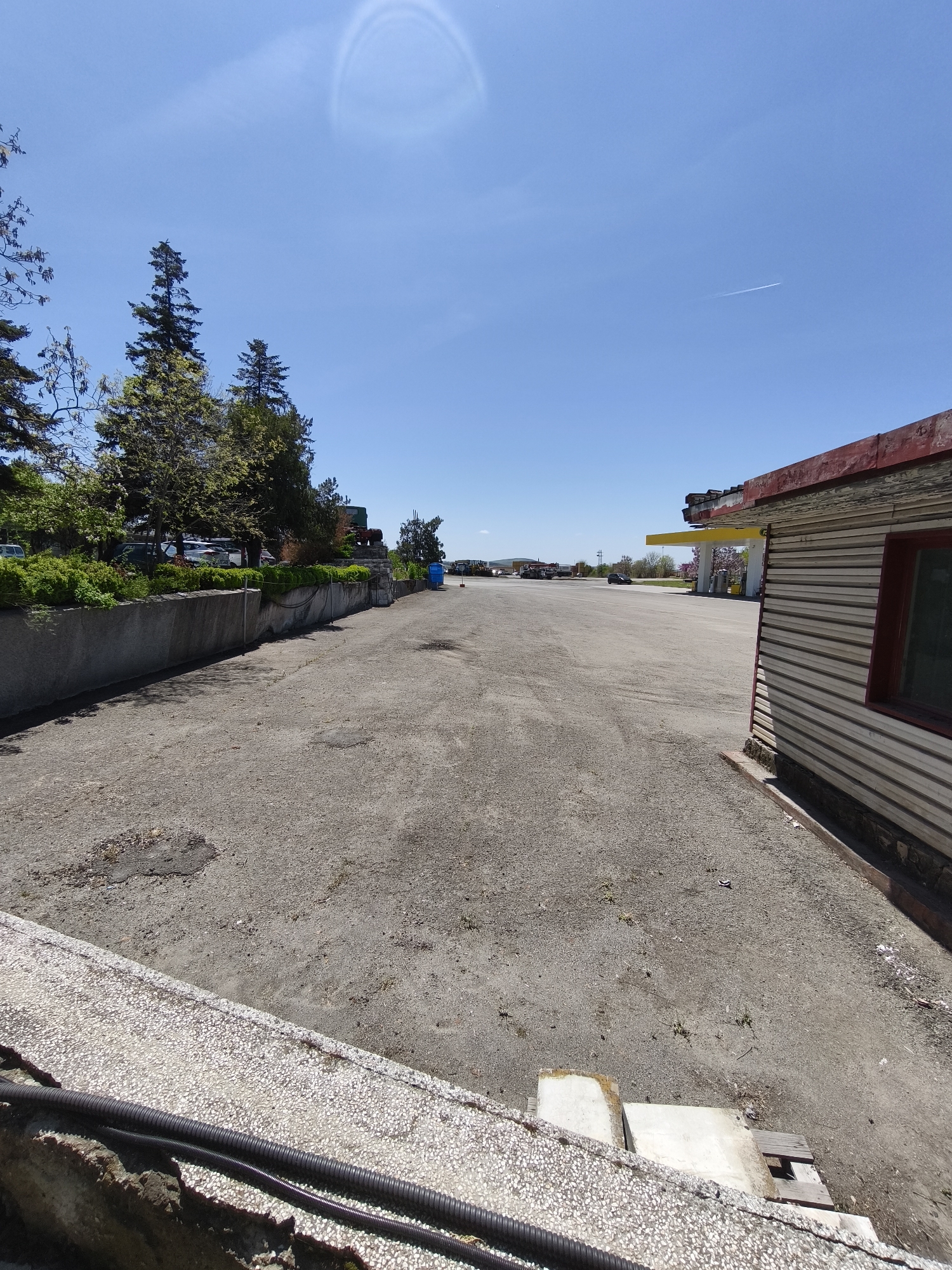 The image captures an expansive, mostly empty asphalt lot under a bright, clear blue sky on a sunny day. The scene appears to be an abandoned or neglected roadside area, possibly a former gas station, located in Burgas, Bulgaria.
In the immediate foreground, a low, textured concrete wall runs horizontally, with black corrugated pipes lying along its base. The ground is rough, cracked asphalt with patches of dirt and sparse vegetation.
To the left, a taller concrete retaining wall supports a bank of lush green bushes and mature trees, mostly evergreen pines and deciduous trees. Several vehicles are partially visible parked behind this foliage. Further down the lot, a blue portable toilet can be seen near the left wall.
To the right, part of a dilapidated single-story building with corrugated metal siding is visible. It features a red-painted roof trim and a window with a dark frame. Beyond this building, a yellow canopy, characteristic of a gas station, extends over what appears to be fuel pumps in the mid-distance. The ground in this area of the lot also shows signs of wear and neglect.
The lot stretches far into the background, where more distant buildings and what look like other parked vehicles are visible. A dark car can be seen driving or parked further down the road. The sky above is a vibrant blue, with a faint airplane contrail stretching across it and a subtle, circular atmospheric effect, possibly a sun halo or lens flare, visible in the upper left. No people are visible, and there is no discernible text in the image.