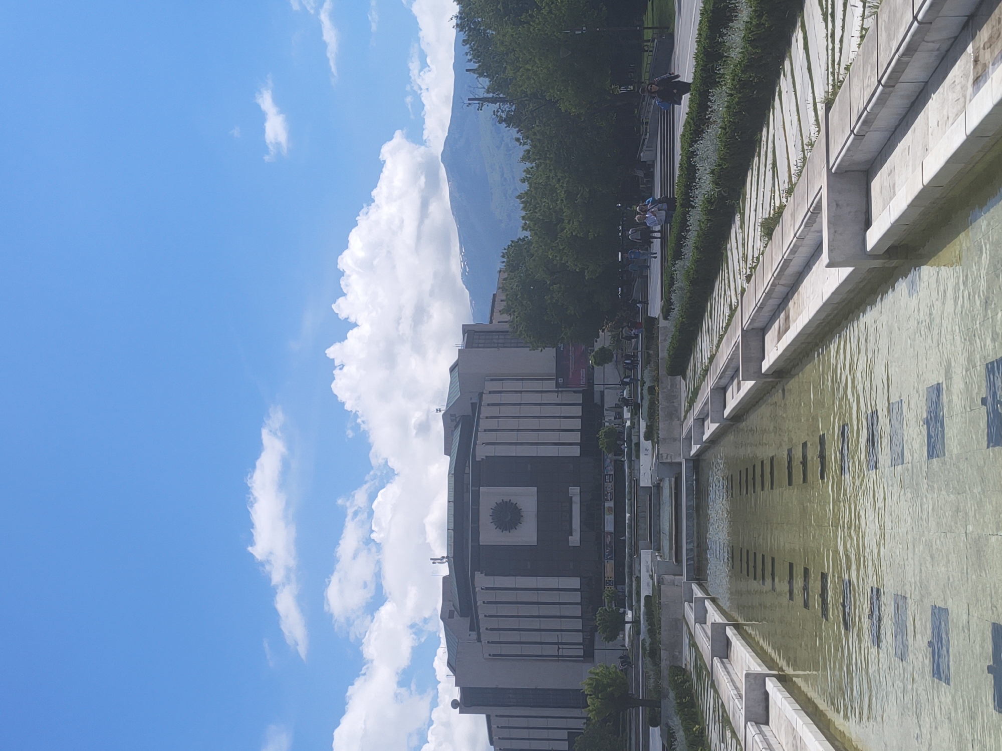The image captures a view in Sofia, Bulgaria, featuring the National Palace of Culture (NDK). The NDK, a large, boxy building with a prominent central clock-like feature, is positioned in the middle ground against a backdrop of a clear blue sky with fluffy white clouds and a mountain range.
In the foreground, a long rectangular pool or water feature reflects the architecture and sky. The pool is flanked by concrete ledges and landscaping.
Along the right side of the pool, a paved pedestrian area is visible, populated with several people walking or standing. Trees line this path.