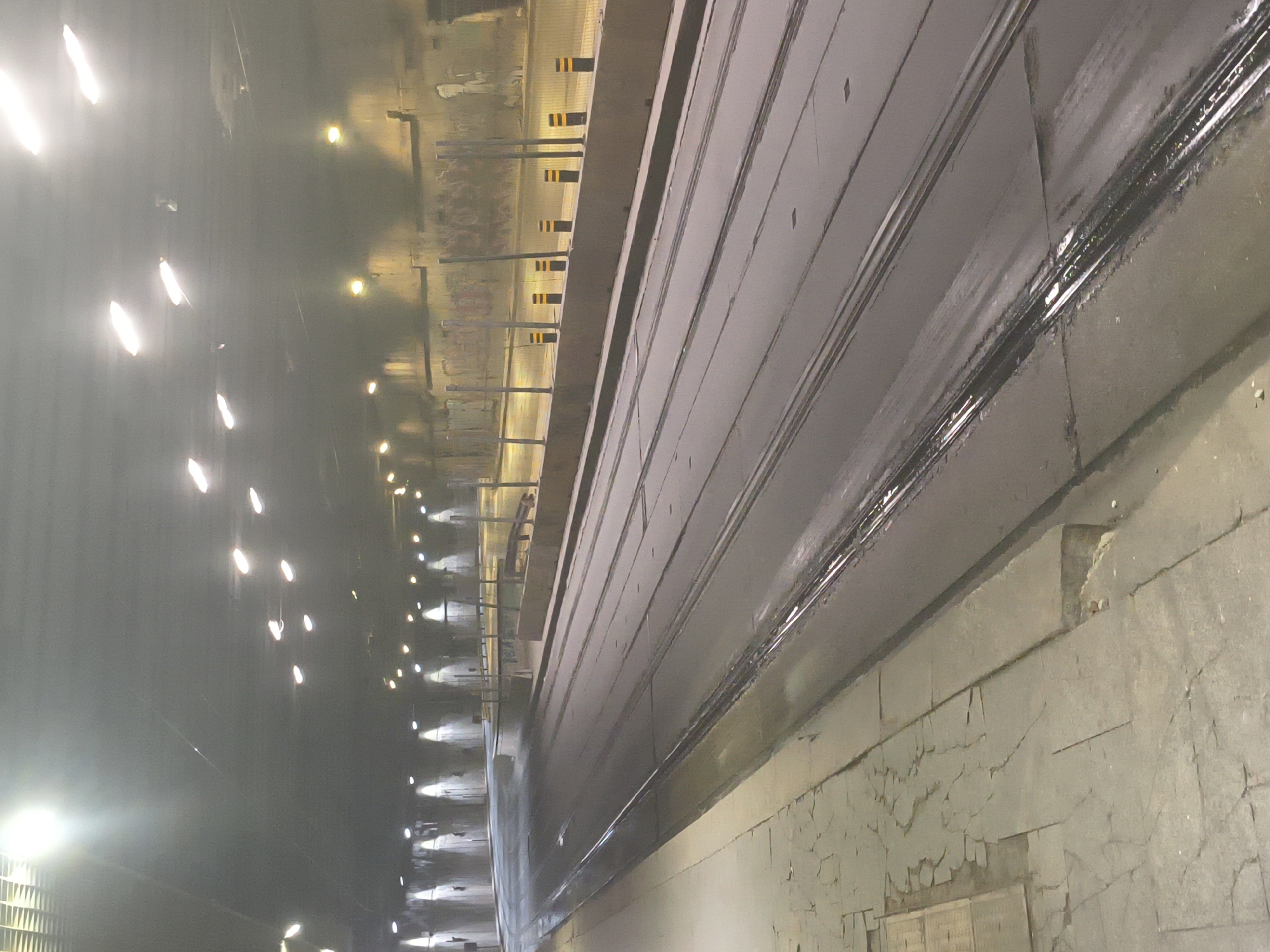 The image depicts a section of the Sofia Metro, Bulgaria. The scene appears to be within a tunnel, evidenced by the repeated pattern of lights lining the tunnel wall. A raised platform or walkway runs along the right side of the image, with the tunnel curving into the distance. Cracks and aged textures are visible on the tunnel walls, indicating age and wear. The perspective and lighting suggest a slightly distorted depth of field, with the foreground appearing closer than it is. No people are visible in the frame.