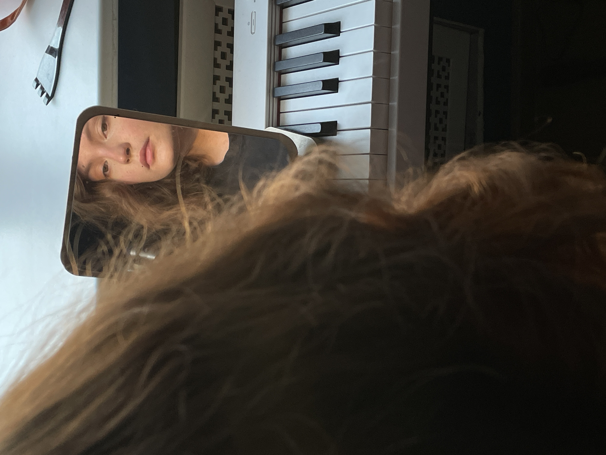 The image shows a person's face reflected in a mirror, with a piano keyboard visible in the background. The mirror is positioned in a way that the viewer also sees the back of the person's head in the foreground, creating a partial obstruction. The reflection reveals a young person with light skin and brown hair. A comb-like object can be seen near the top left of the frame. The location is not discernible from the given image.