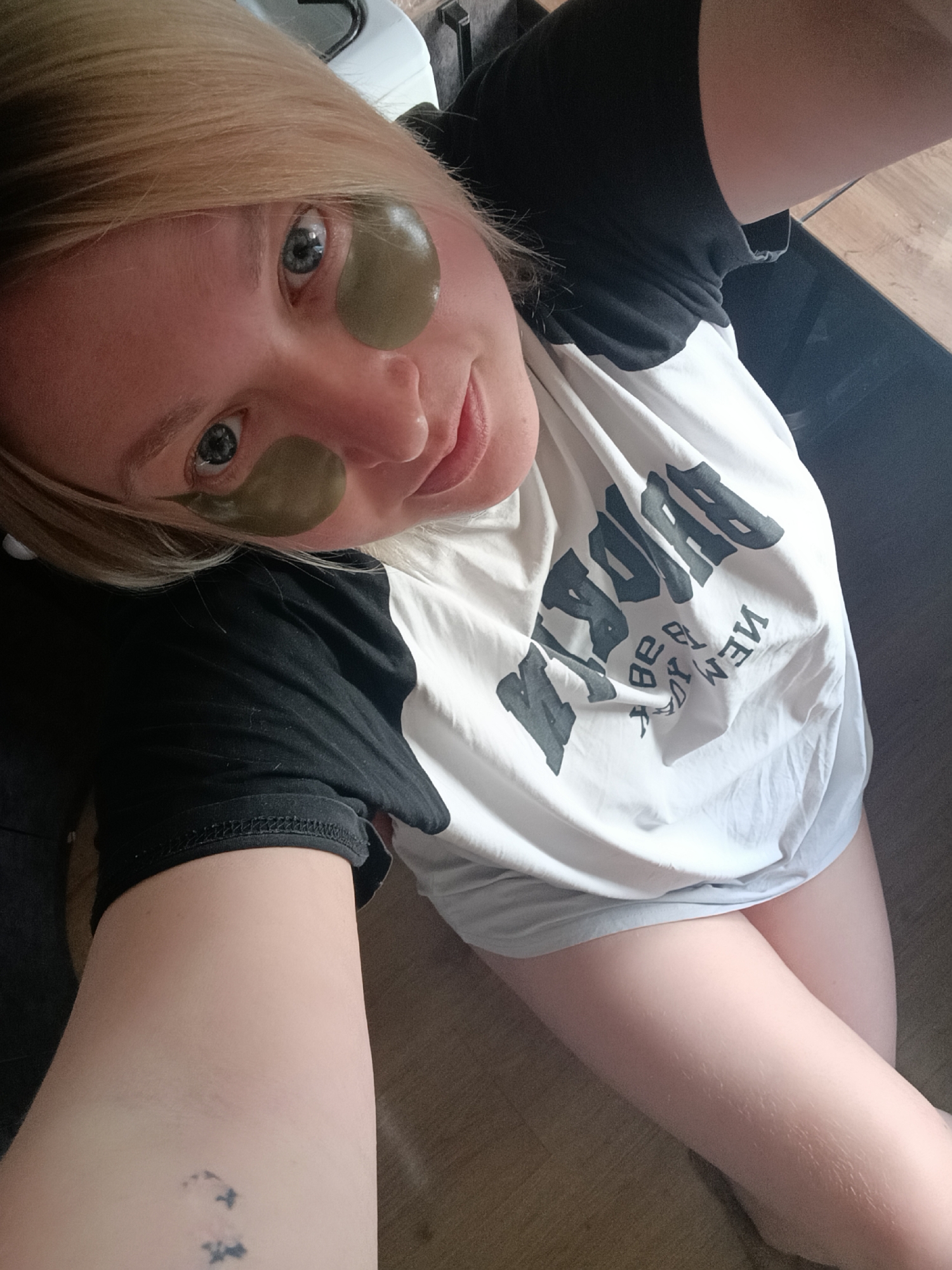 A young woman with blonde hair and striking blue eyes is depicted in a close-up, high-angle selfie taken indoors, likely in Chaikovsky, Russia. She looks directly at the camera with a soft, pleasant smile. She is wearing green, gel-like under-eye patches, suggesting a skincare routine or a moment of relaxation.
She is dressed in a casual t-shirt with black sleeves and a white body. Mirrored text is visible on the front of her shirt, clearly spelling "BROOKLYN" on the upper line and "NEW YORK" below it, rendered in large, bold black letters. A small, dark-colored, abstract tattoo is visible on her inner left forearm, which is extended towards the camera. Her fair-skinned legs are partially visible, resting on a light-colored surface that appears to be a wooden floor. The background is softly blurred, showing glimpses of wooden textures and dark furniture, indicating a home environment. The scene is illuminated by natural light, suggesting it is daytime. The woman's expression conveys a sense of calm and contentment.