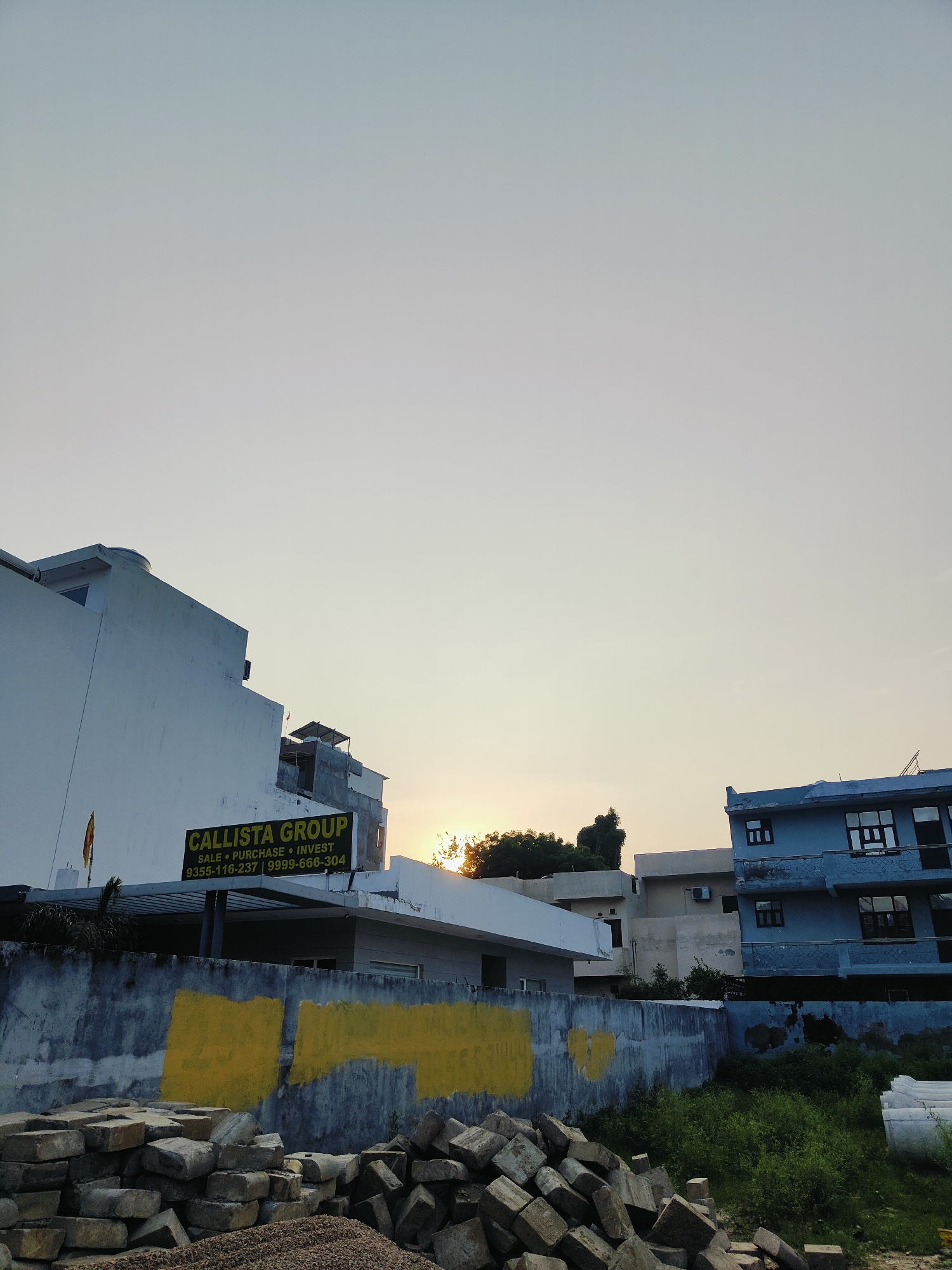 The image depicts an outdoor scene, likely in Gurgaon, India, at dusk. In the foreground, a pile of construction debris, consisting of broken bricks and gravel, is visible. Behind the debris is a concrete wall with large, roughly painted yellow rectangular shapes.
Further back, several buildings are present. On the left, a white building with a prominent sign that reads "CALLISTA GROUP SALE • PURCHASE • INVEST 9355-116-237 | 9999-666-304" is partially visible. Adjacent to it, a multi-story building painted in a pale blue hue with dark-framed windows is situated on the right. The sky is a pale, hazy yellow, suggesting either a sunset or sunrise, with the sun's rays peeking through trees behind the buildings. The environment appears to be an urban or semi-urban area undergoing some form of construction or renovation, given the debris and the presence of real estate signage. No people are visible in the frame.