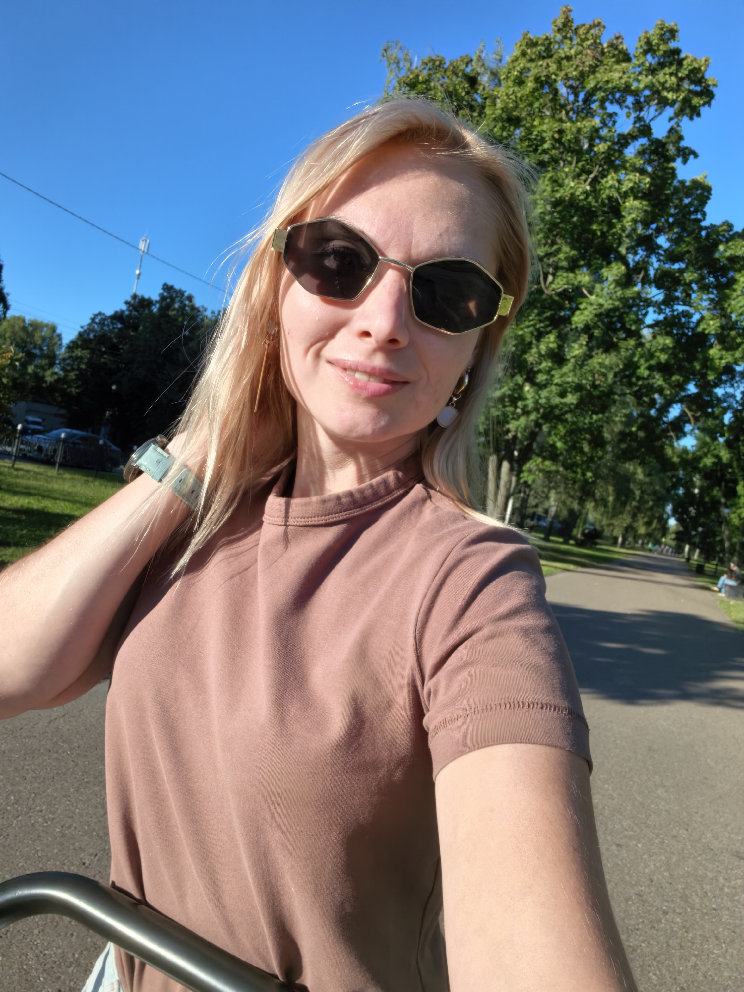 A selfie is taken by a blonde woman wearing sunglasses and a pinkish-brown t-shirt. She has a light skin tone and is smiling slightly. The background is a park with a paved path, green trees, and a clear blue sky. The scene appears to be outdoors during the daytime.