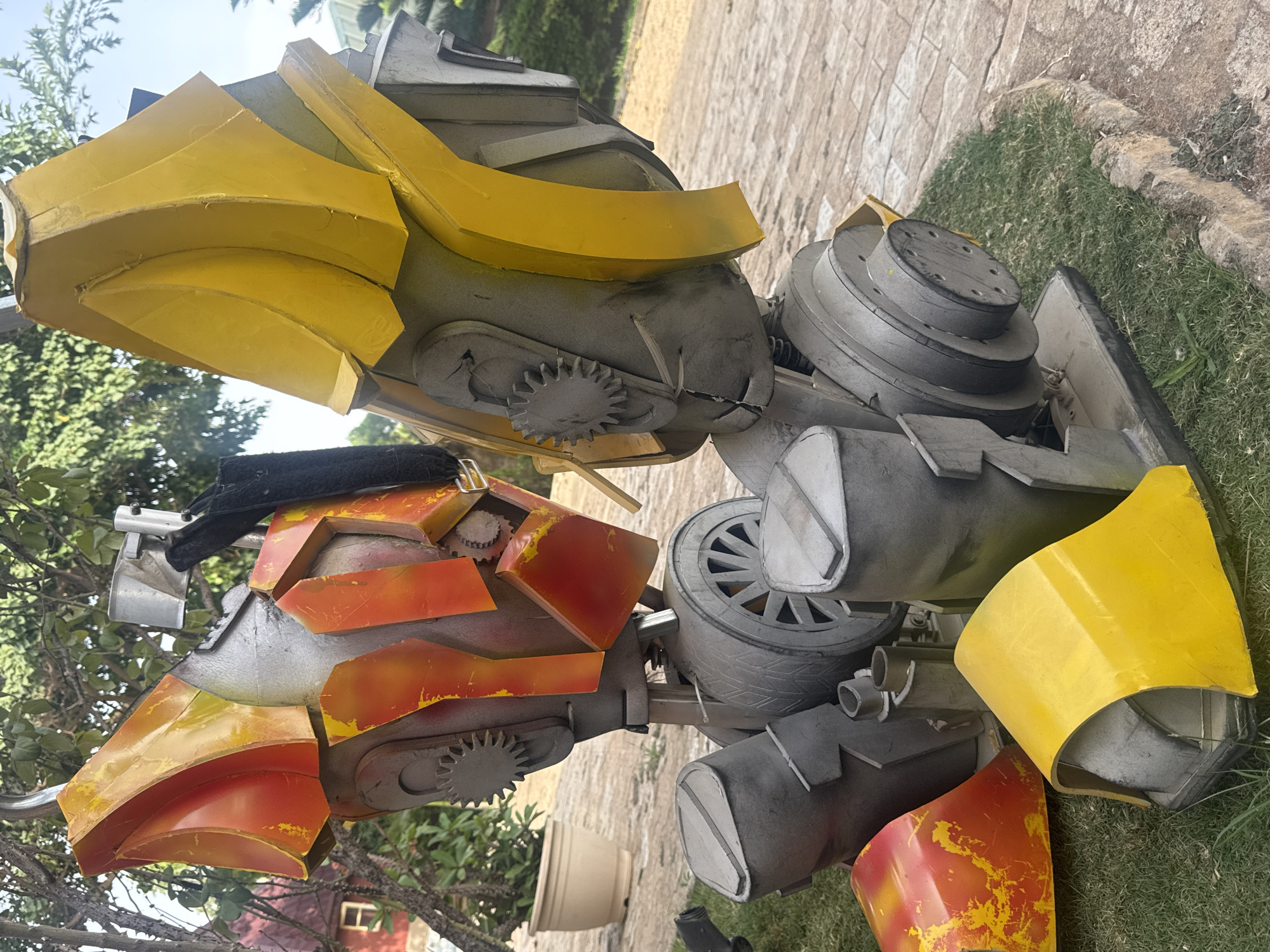 The image shows a large, homemade mechanical sculpture on a grassy area. The sculpture is composed of various materials, including metal and painted foam. The upper portion is primarily yellow and gray, while the lower portion features red and orange elements. The sculpture incorporates gears and wheel-like structures. The background shows trees, a building, and a paved area. There is no indication of the sculpture's location being Jos, Nigeria.