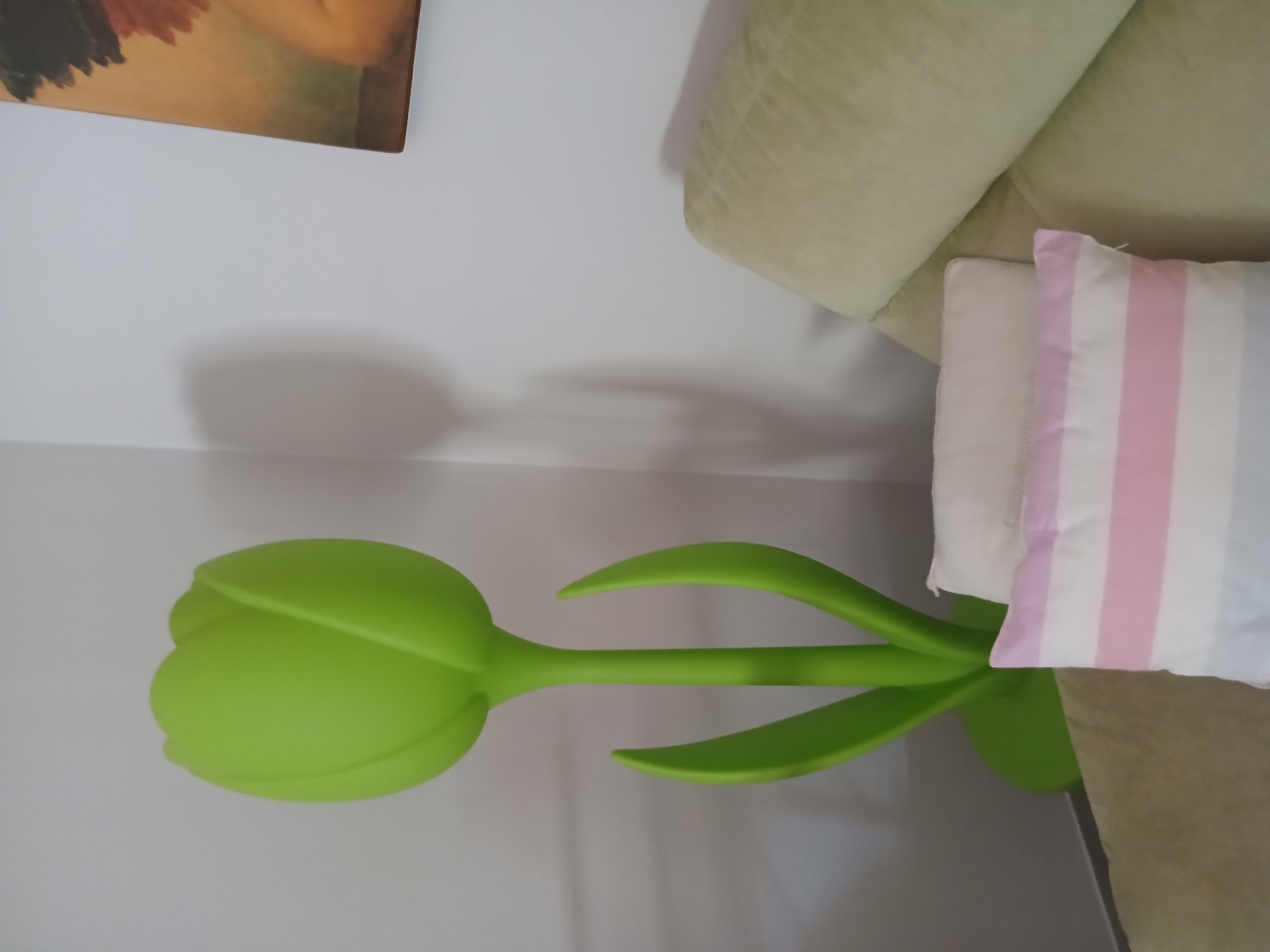 The image captures an indoor scene featuring a lime green tulip-shaped floor lamp standing on a circular base. The lamp is positioned to the left of the frame. To the right, a portion of a sofa is visible, adorned with two pillows: one white and one with horizontal stripes in pink, white, and light blue. The wall behind the lamp shows a subtle shadow cast by the object, and a painting with warm tones, possibly depicting a figure, hangs on the wall in the upper right corner. The overall setting suggests a domestic interior, possibly a living room. This image was taken in Monsummano Terme, Italy.